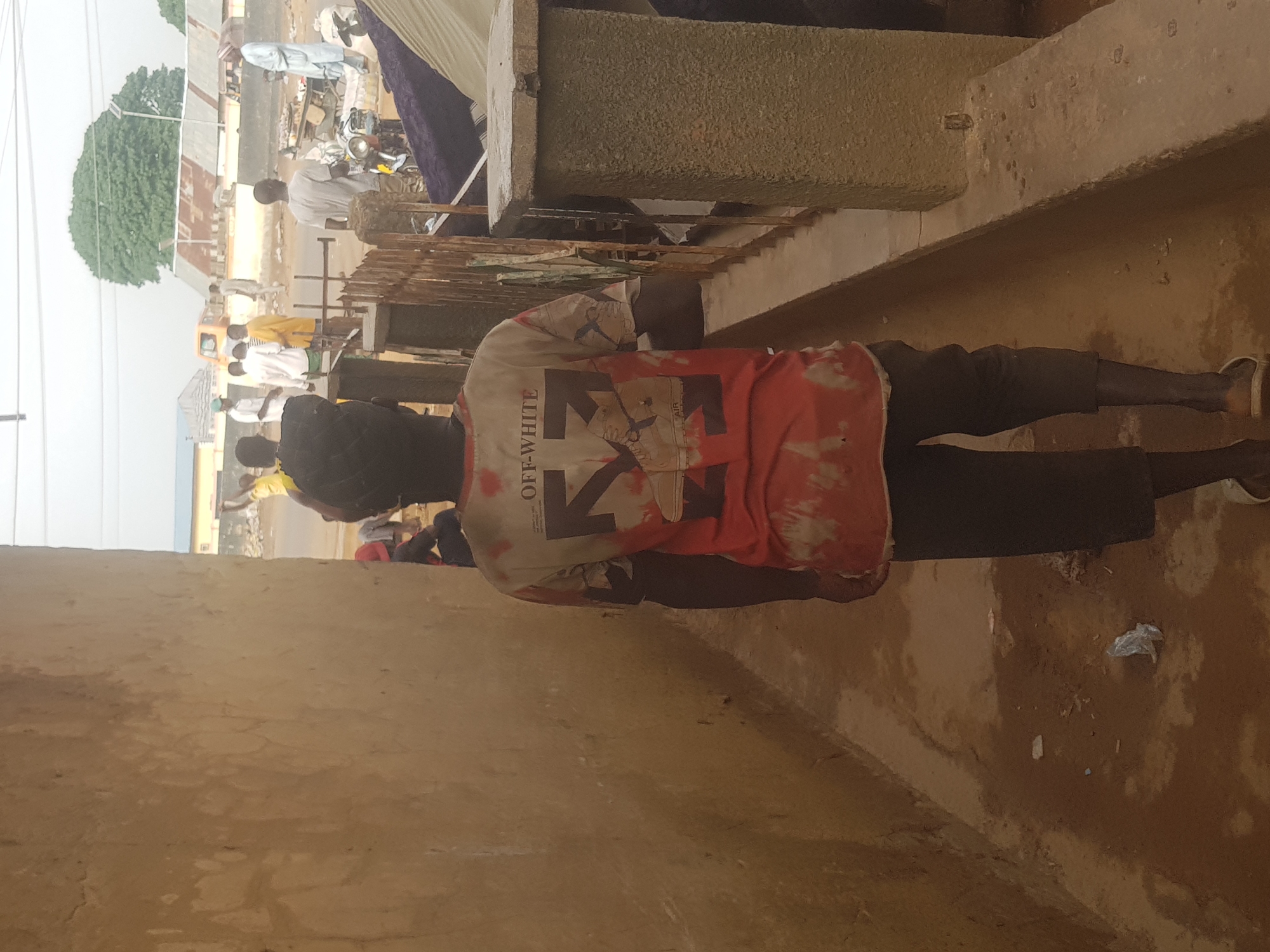 A street in Kufena, Nigeria, shows a man in a red and white "OFF-WHITE" shirt walking on a paved walkway. People are visible in the background on the street, alongside cars, mopeds, and structures. A large tree is in the distance.