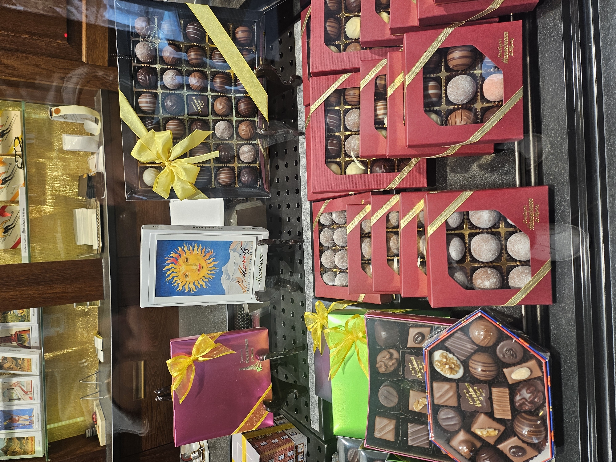 This image captures a display of assorted chocolates inside a confectionery shop. The chocolates are arranged in various boxes, some clear and some red, all adorned with yellow ribbons. The confectionery's name, "Confiserie Hanselmann St. Moritz," is visible on some of the packaging, indicating the location as St. Moritz, Switzerland. The display is set against a dark background, with a mirrored surface reflecting the scene. In the background, shelves are visible holding more items, possibly other confectioneries or related products. The lighting is bright, highlighting the chocolates and their packaging. There are no people visible in the image. The overall impression is of a well-organized and appealing product display.