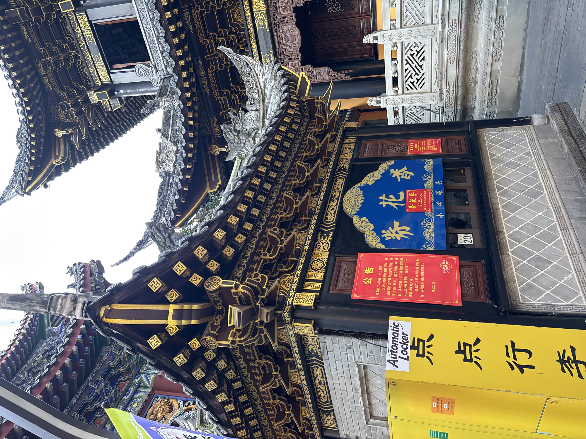 The image shows a traditional Chinese building in Yuzhong District, China. The architecture features intricate dark wood and gold detailing, with a multi-tiered roof. Signs in Chinese are visible on the building, including a notice about reducing incense pollution and offering free incense. A yellow "Automatic Locker" sign is present. The overall setting is urban.