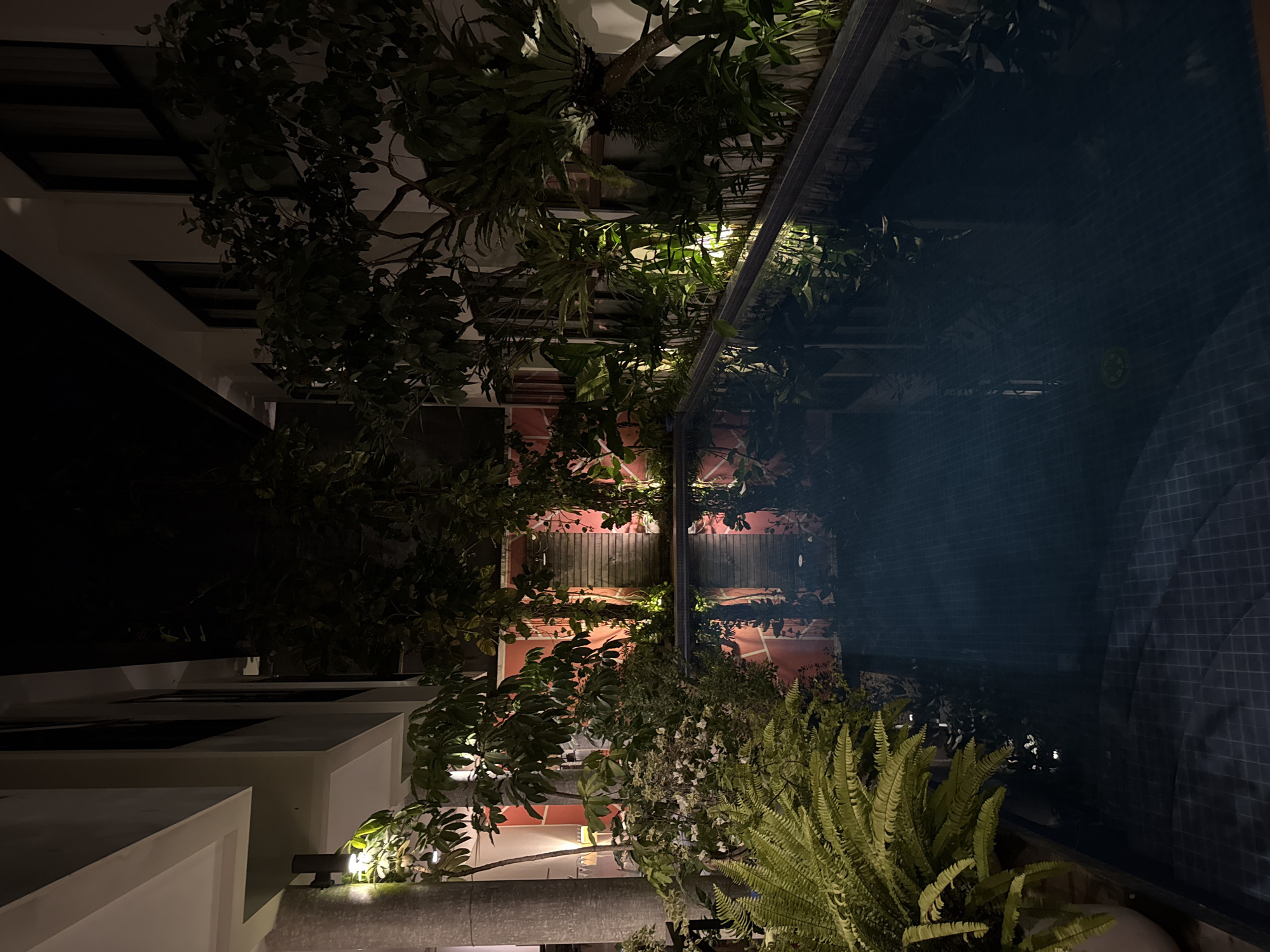 This is an exterior nighttime shot, likely from a higher vantage point, in North Kuta, Indonesia. A dark, tiled pool dominates the right side of the image, reflecting the surrounding architecture and vegetation. On the left, a building with light-colored walls and what appear to be open-air spaces is visible, interspersed with tropical foliage. Additional structures featuring reddish-toned walls are seen between the greenery. The scene is illuminated by artificial lighting. No people are visible.