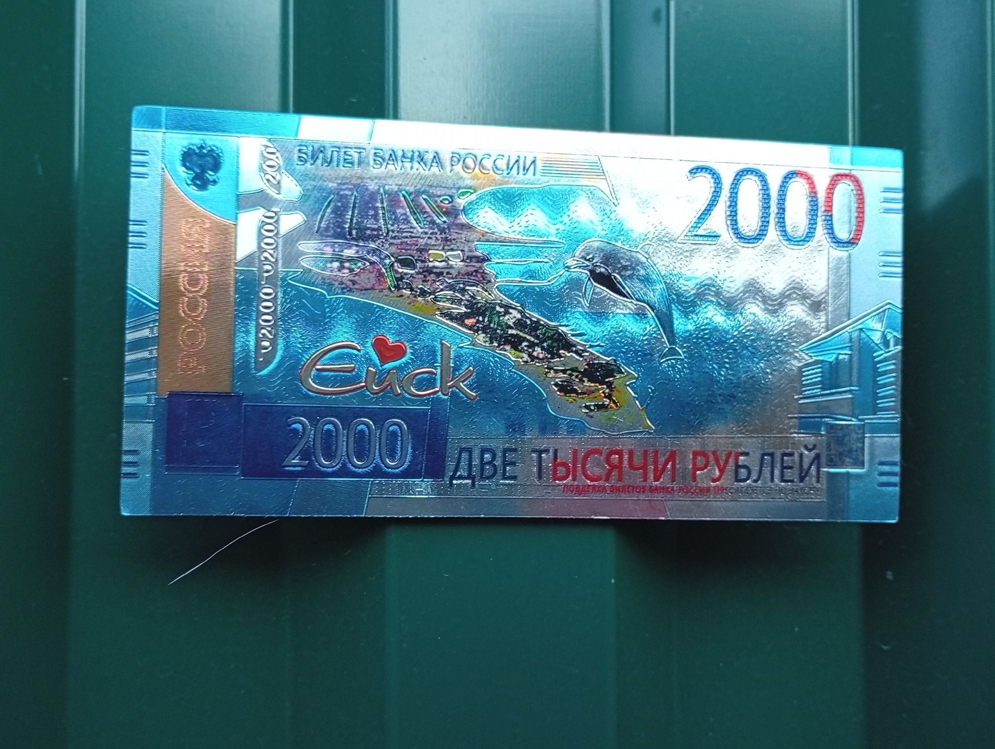 The image shows a close-up view of a Russian 2000 ruble banknote. The banknote features a holographic design with a dolphin and a ship. The text on the banknote is in Russian and includes "БИЛЕТ БАНКА РОССИИ" (Bank of Russia ticket) and "ДВЕ ТЫСЯЧИ РУБЛЕЙ" (Two thousand rubles). The banknote is placed on a dark green textured surface, possibly a metal fence or corrugated material. There is no indication of the location being Pavlovsky Posad, Russia.