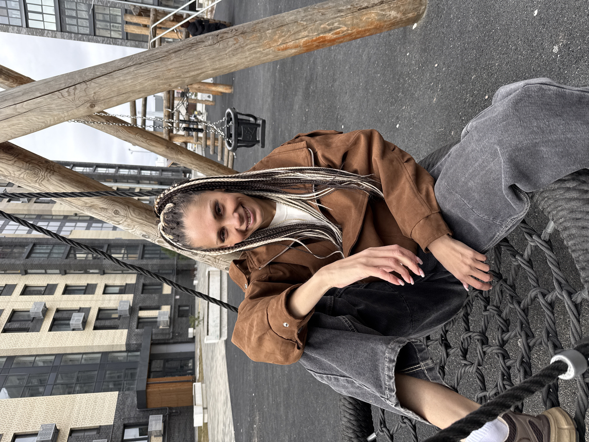 A young woman with braided hair is sitting in a rope swing structure in what appears to be an urban park. She is wearing a brown jacket over a white top, black pants, and light gray denim overalls. The swing is suspended from wooden support beams. In the background is a modern apartment building constructed of brick. The location appears to be the city of Rusanovo, Russia.
