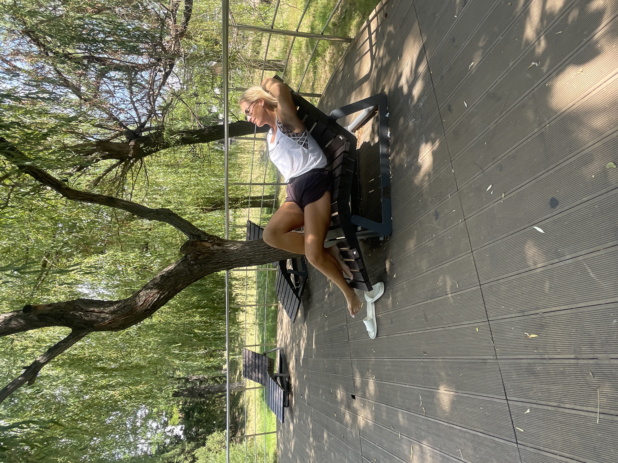 A blonde woman is relaxing on a dark wooden deck in Novorossiysk, Russia. She is lying on a dark recliner chair, wearing a white tank top and dark shorts. Her eyes are closed, and she appears to be enjoying the sunshine. The deck is surrounded by lush green trees, and there are other recliner chairs scattered around the area. The scene suggests a peaceful and relaxing outdoor setting.