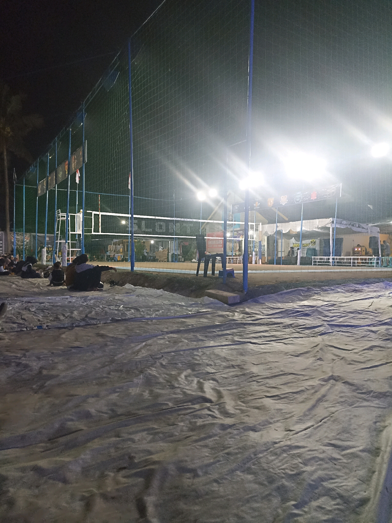 The image depicts an outdoor volleyball court at night in Pacitan, Indonesia, illuminated by bright floodlights. A net is stretched across the court, with volleyball posts on either side. Spectators are seated on the ground in the foreground, some appearing to be watching the game or an event. Behind the court, there are tents and structures, possibly for vendors or participants. Flags are visible, and banners with text are hung along the fence. The ground is covered with tarps in the foreground, suggesting preparation or a temporary setup.