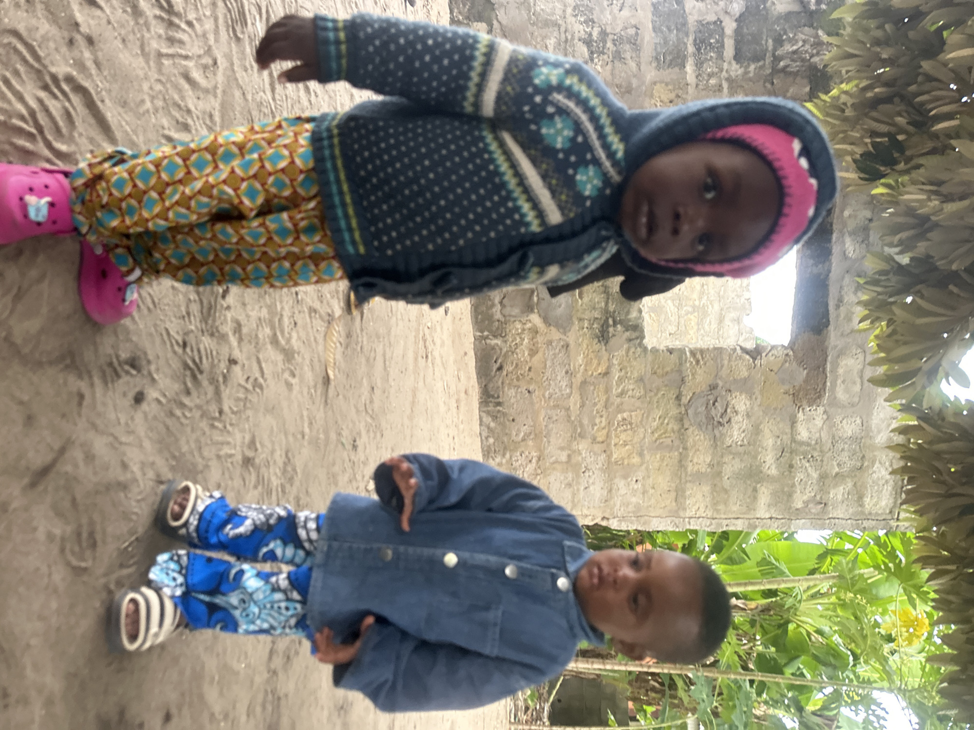 In Badagry, Nigeria, two young children stand on a sandy ground in front of a weathered, partially constructed brick building. The child on the left, wearing a patterned blue sweater with a pink-lined hood and vibrant patterned pants, has their hands near their waist. The child on the right is dressed in a denim jacket and blue patterned pants, with their hands held out to the sides. In the background, lush green foliage and trees are visible, suggesting an outdoor setting. The sunlight casts a soft light, illuminating the scene.