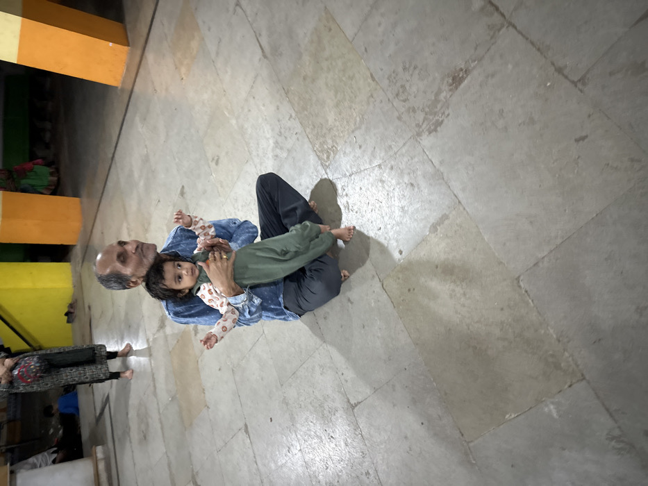 An older man sits on the floor with a baby lying across his legs. The man is wearing a blue denim shirt and black pants. The baby wears a patterned long-sleeved shirt and green pants. They are both barefoot. They are on a stone-tiled floor. In the background, there are blurred figures of people walking by. A section of a colorful building structure is also partially visible.