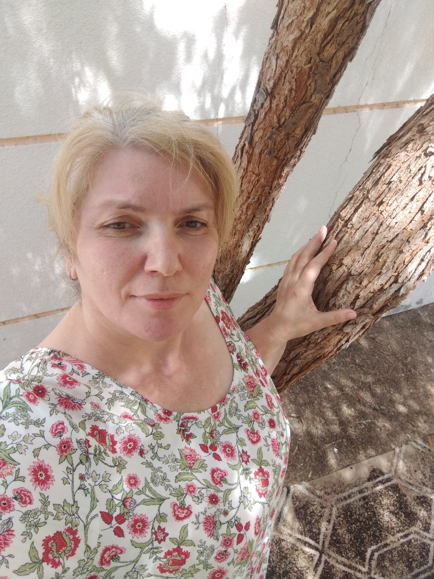 A middle-aged woman with short blonde hair and fair skin stands outdoors. She wears a floral-patterned top. She is touching a tree trunk with her right hand. The tree trunk appears to have rough, textured bark. The background consists of a white wall with some shadow patterns and a ground area with a textured pattern. The location is Al Mas'oudi, United Arab Emirates.