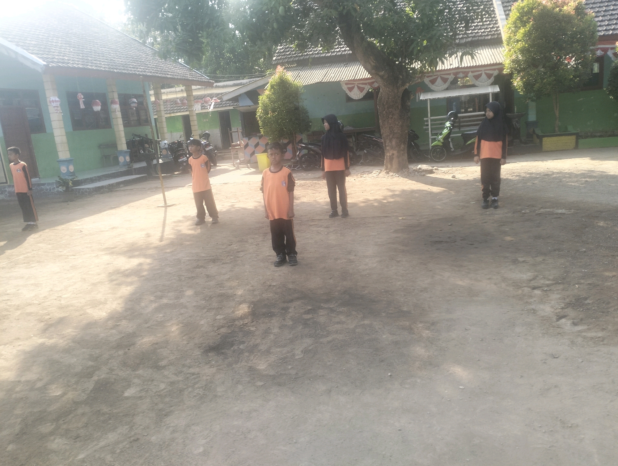 The image depicts an outdoor scene in Blabakan, Indonesia, likely a schoolyard. Four children, dressed in orange shirts and dark pants, are standing on a dirt ground. Two of the children are boys, and two are girls wearing headscarves. In the background, there are two school buildings with tiled roofs, a tree, and some motorcycles. The area appears to be a rural or semi-rural setting. The sun is bright, casting shadows on the ground.