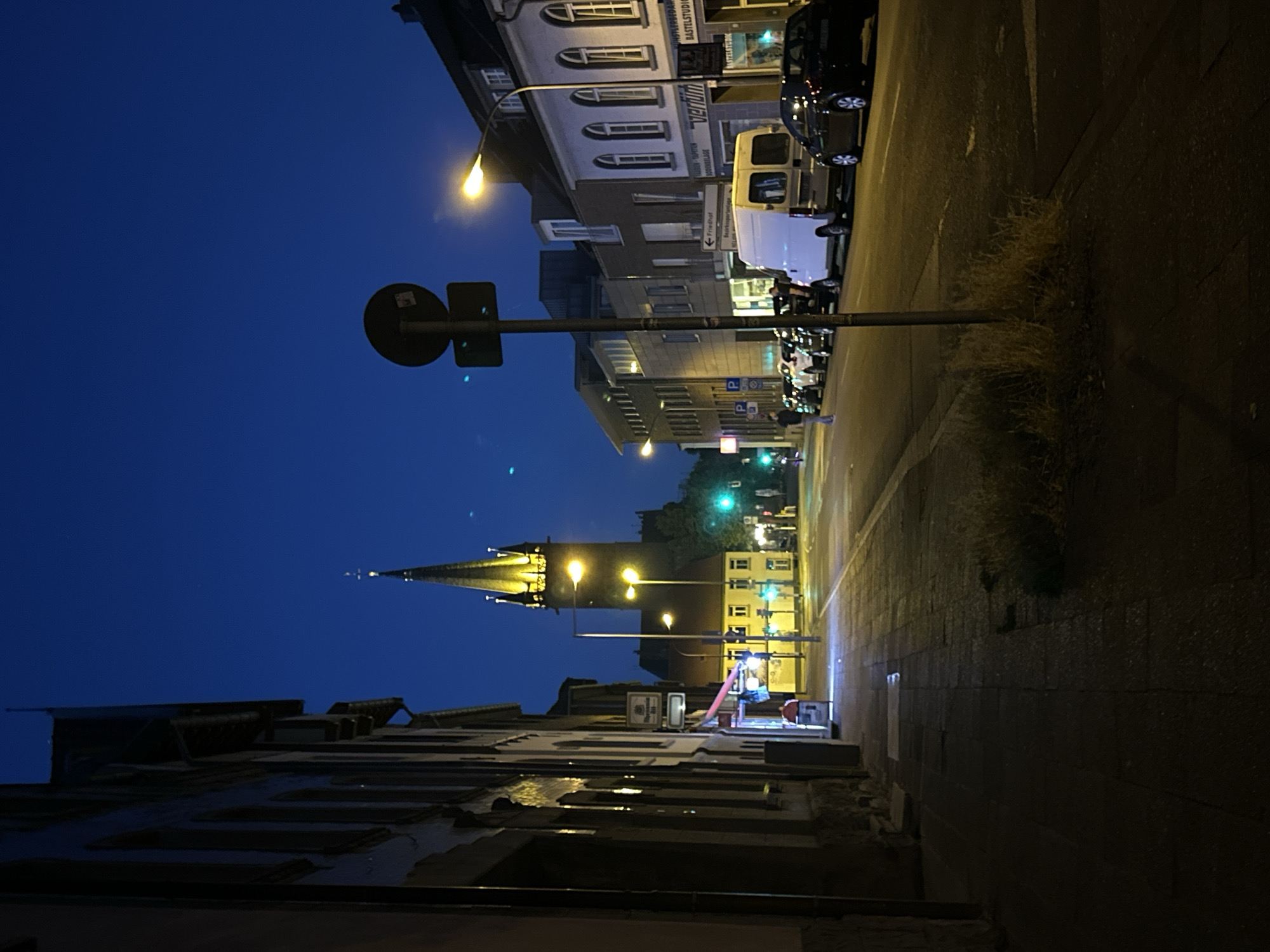 The image captures a street scene in Krefeld, Germany, at dusk or early night. A tall church spire, illuminated with yellow lights, dominates the background. The street is lined with buildings on both sides, featuring storefronts and residential windows. Parked cars, including a white van and several passenger vehicles, are visible along the curb. Streetlights cast a warm glow on the asphalt and sidewalk. A lone figure walks on the sidewalk on the left side of the street. The sky is a deep blue, indicating the transition from day to night.
