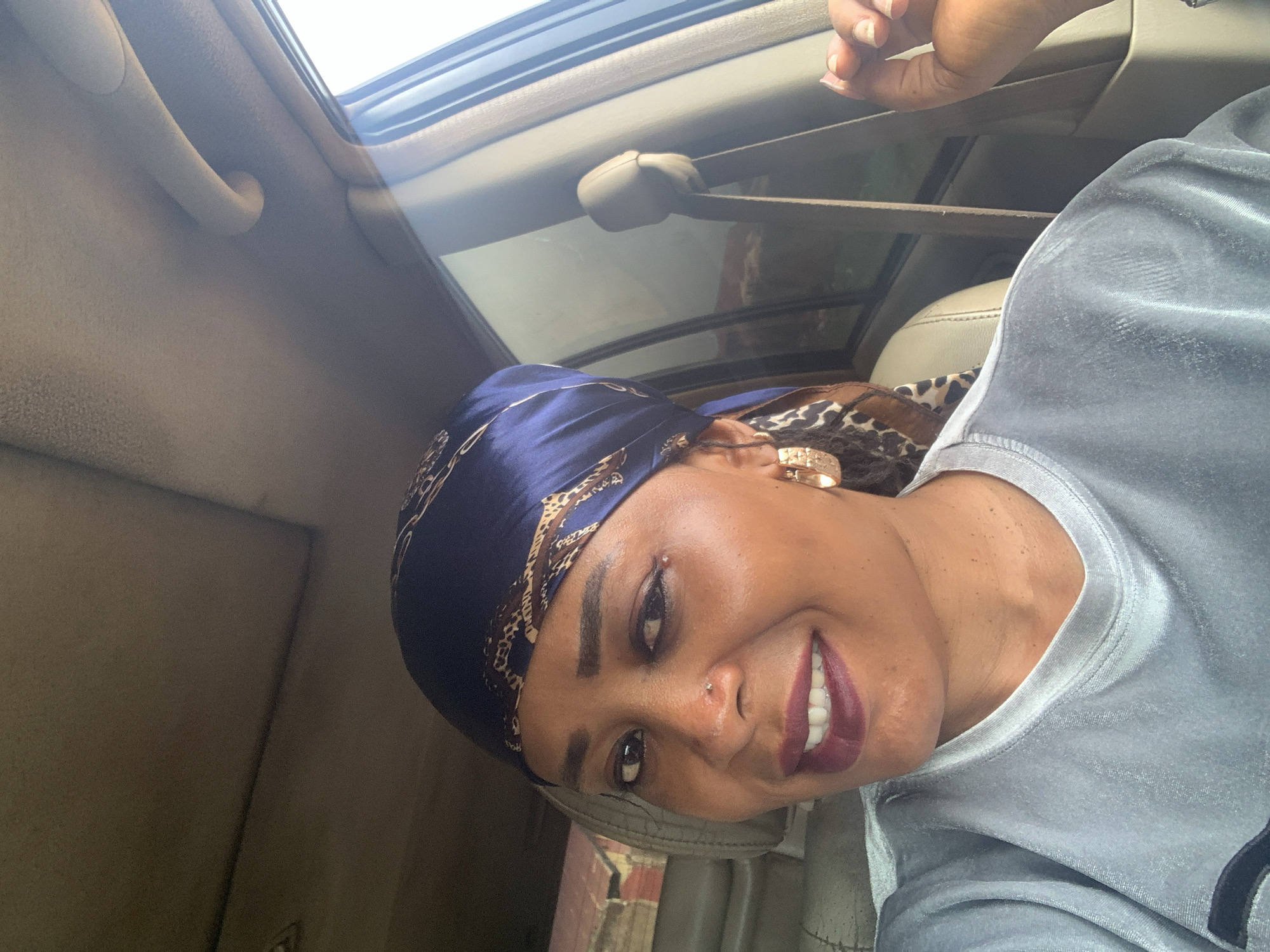 A woman wearing a blue headscarf and a grey shirt is positioned inside a car with a sunroof. She is smiling. The car's interior includes a seatbelt and an overhead handle. Based on the image's context, the location is presumed to be Sabon Tasha, Nigeria.