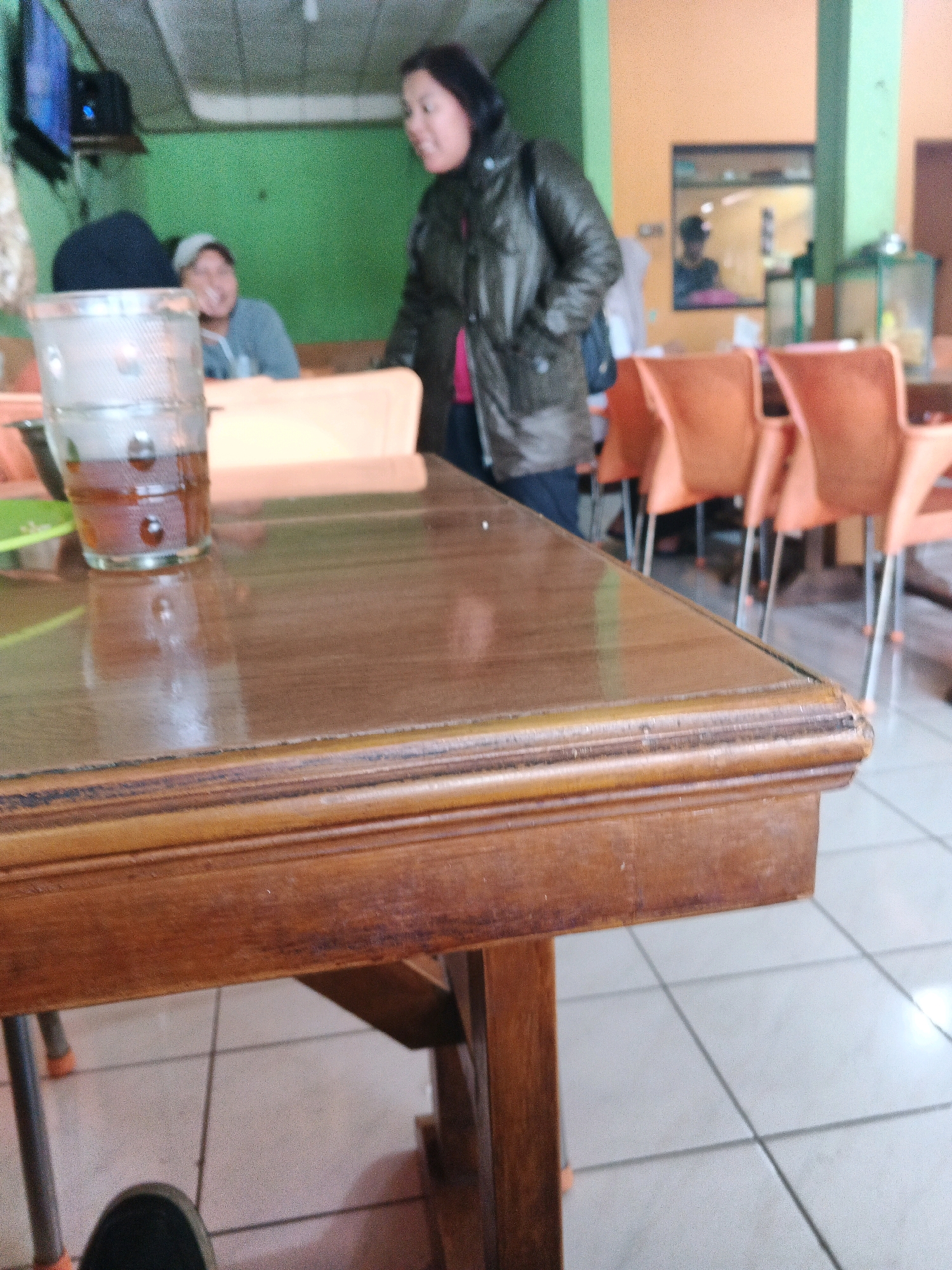 A medium-shot, outdoor photograph captures a wooden table with a glass of iced tea in the foreground, with several people and orange chairs blurred in the background. The table is made of dark wood and appears to be well-worn. The glass of iced tea is a tall, cylindrical glass filled with a dark amber liquid and ice cubes. The background shows three people and several orange chairs. A woman in a dark jacket and pink shirt stands on the right, facing away from the camera. A man in a grey shirt and a cap is seated to her left, smiling. Another person is partially visible to the far left. The scene appears to be a cafe or restaurant in Ungaran, Indonesia, with a green wall and an orange pillar visible in the background. The overall lighting is dim, suggesting it is an indoor setting with artificial lighting.