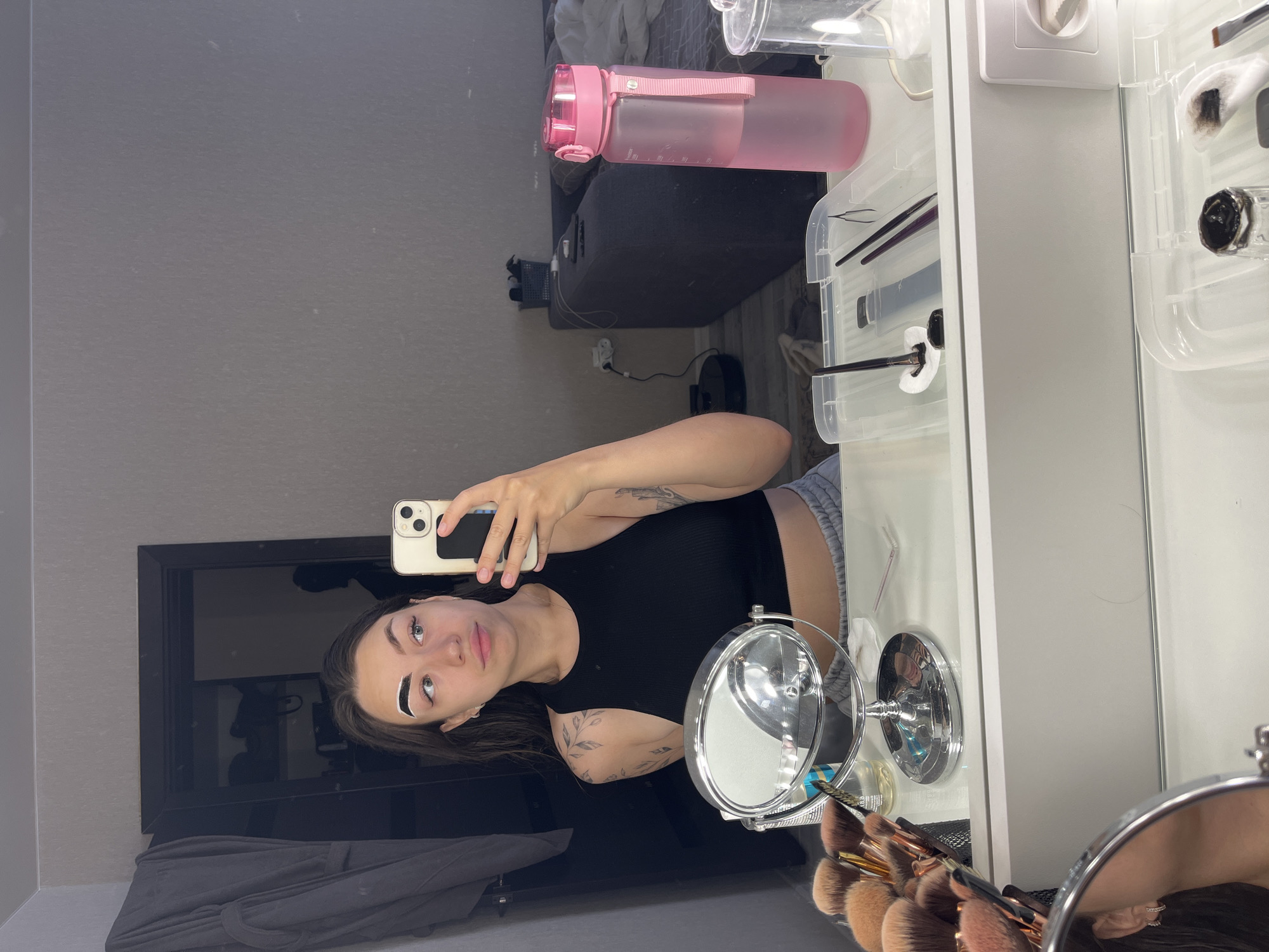 In a room in Chaikovsky, Russia, a young woman with dark hair and tattoos on her arm is taking a selfie in a mirror. She is wearing a black tank top and grey sweatpants, and her left eyebrow is covered in white product, likely for brow lamination. She is holding a white smartphone in her right hand, and her left hand is holding a makeup brush.
In the foreground, a vanity table is visible, with various makeup brushes, a magnifying mirror, and a clear plastic container holding makeup tools. A pink water bottle is on the right side of the frame, and a sofa is visible in the background. The room appears to be a bedroom or dressing area, with a closet and a doorway leading to another room.