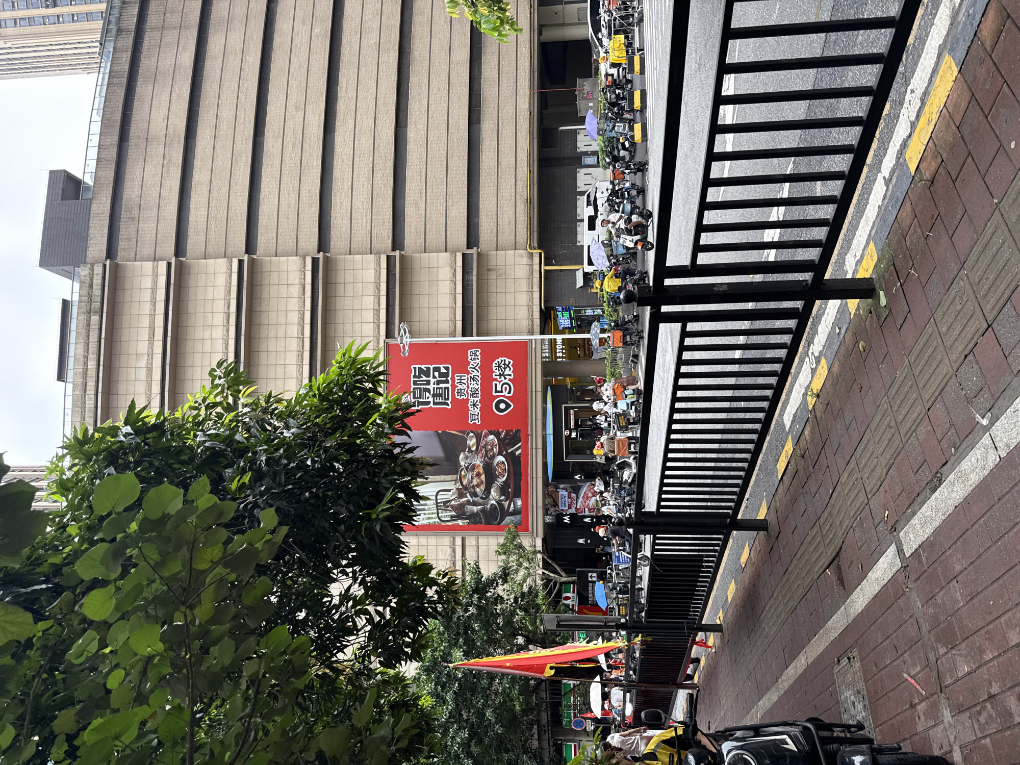 A vertical, eye-level shot captures a street scene in China's Haizhu District, dominated by a tall, modern building with a textured beige facade. In front of the building, a large red banner advertises "Tang Ji Guizhou Dou Mi Sour Soup Hot Pot" and indicates it is on the 5th floor. The banner features an image of a steaming hot pot meal.
To the right of the banner, a black metal fence separates the sidewalk from the street. The sidewalk is paved with red bricks and marked with yellow and white lines, indicating a crosswalk. Beyond the fence, numerous scooters and motorcycles are parked along the street, some adorned with colorful umbrellas. A few people are visible near the vehicles, engaged in various activities. A white delivery truck is parked amongst the scooters.
The scene is set during the day, under an overcast sky, suggested by the diffused light. Lush green trees partially obscure the view of the building and banner, adding a touch of nature to the urban landscape. The overall atmosphere appears to be a bustling commercial area with significant foot traffic, likely related to food and transportation. The text on the banner suggests a commercial establishment, and the abundance of scooters implies a busy urban environment. The presence of a subway station is also indicated by the text "万能国地铁站".