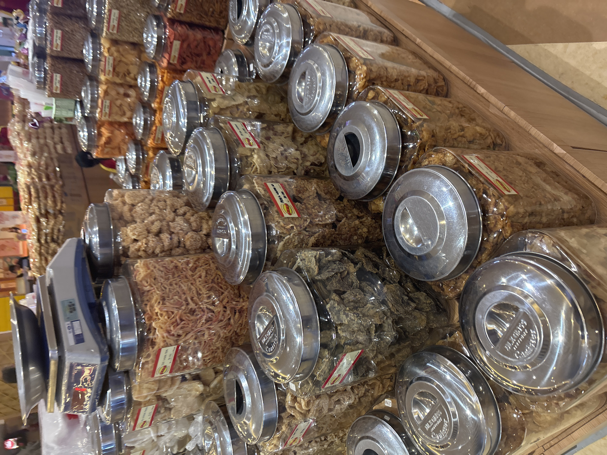 The image depicts a retail display of bulk snacks in clear plastic jars with metallic lids. The jars are arranged on wooden shelves. Several jars are filled with dried, shredded food items, while others contain puffed or crispy snacks. A digital scale with a stainless steel pan is visible in the upper left corner, placed on top of a stack of what appear to be product packaging. In the background, blurred figures of people can be seen, suggesting a bustling market or store environment. The overall impression is a well-stocked snack section of a retail establishment. This scene is consistent with the interior of a supermarket or specialty food store in North Jakarta, Indonesia, where such bulk snack displays are common.