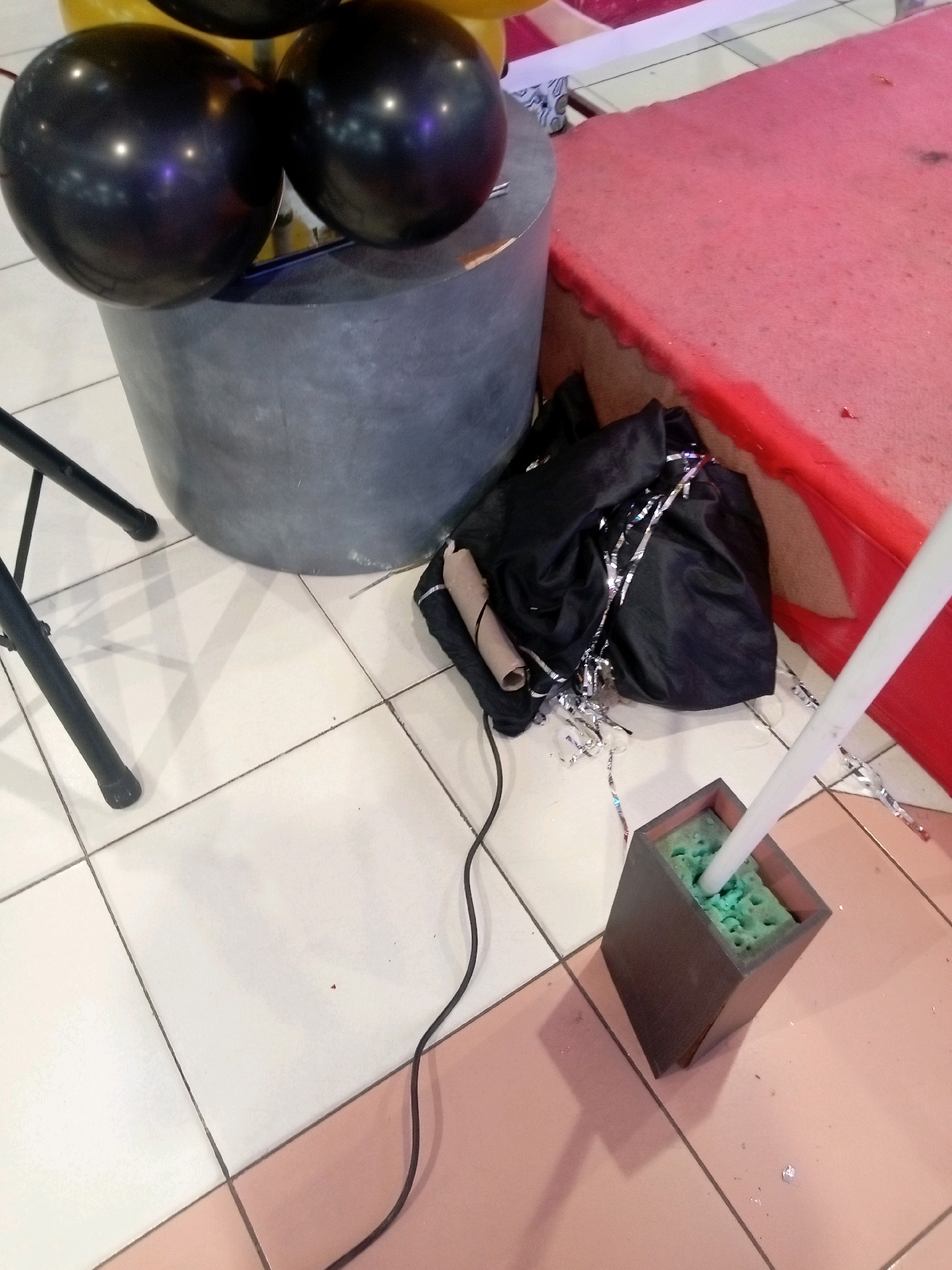 The image depicts an indoor scene, likely a party or event setup, on a tiled floor. In the foreground, a dark wooden planter box filled with green floral foam holds a white cylindrical object. To the left, a black cloth bag is partially covered in silver streamers and a brown cardboard tube. Behind the bag, a large, dark gray cylindrical object, possibly a prop or furniture, is partially visible, topped with two black balloons. To the right of the planter box, a red carpeted platform rises slightly. The lighting suggests an artificial indoor environment. The image does not provide any specific visual cues to determine its location in Batam, Indonesia, or any information about people or actions occurring.