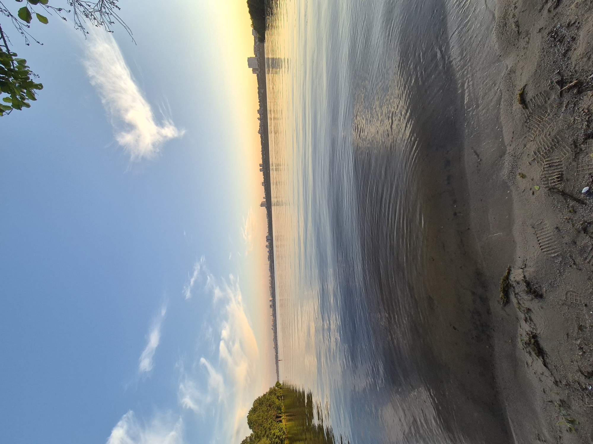 A wide, low-angle shot captures a tranquil lake at sunset. The water surface is calm with gentle ripples, reflecting the soft colors of the sky. The sky is a gradient of deep blue at the top transitioning to warm hues of orange and yellow near the horizon. Wispy white clouds are scattered across the sky. On the left, a lush green tree line borders the water, while a distant cityscape with buildings is visible on the far shore. The foreground features a sandy beach with some scattered debris and footprints. The overall atmosphere is peaceful and serene.