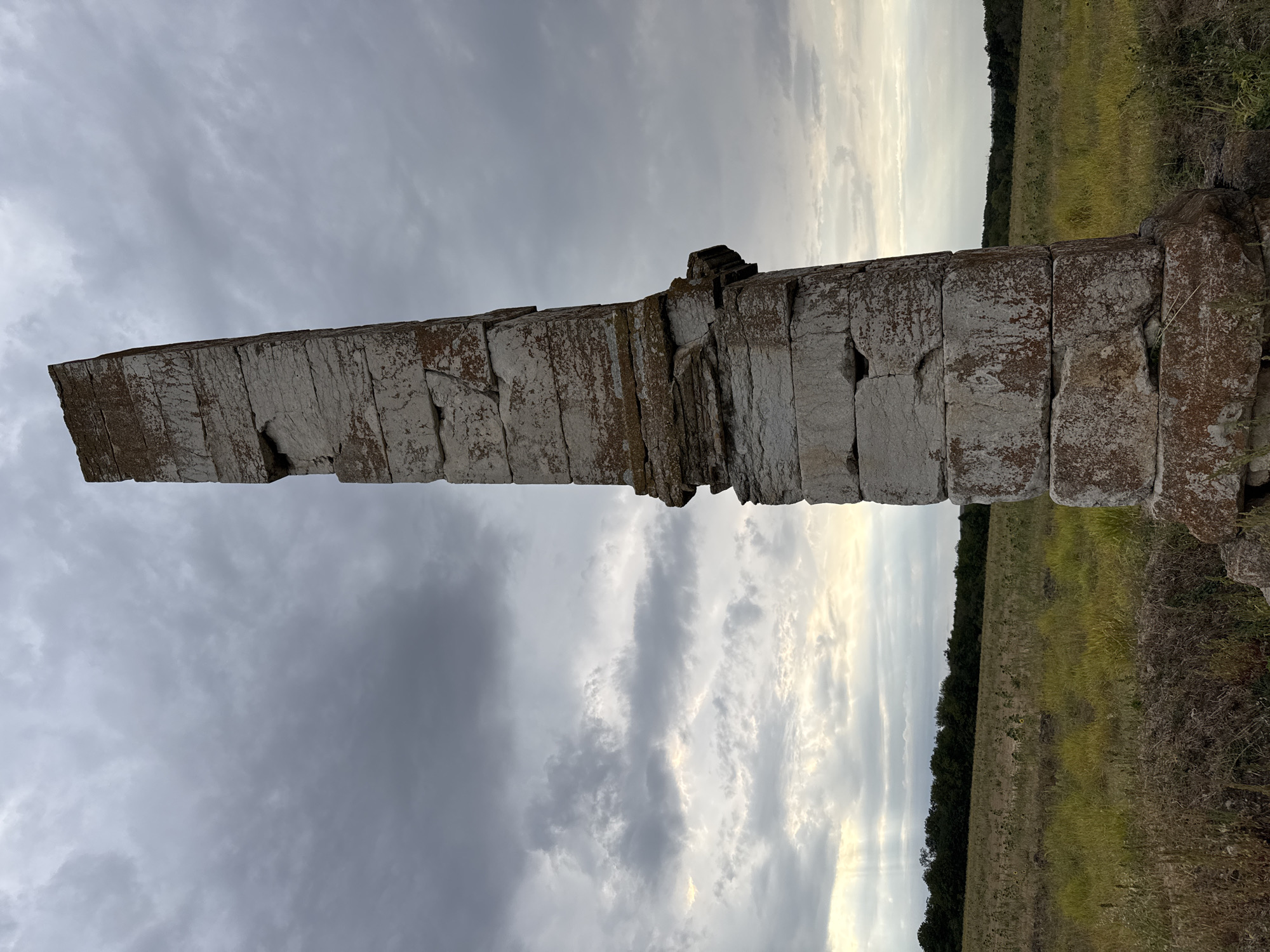 In the city of Lesicheri, Bulgaria, a weathered stone monument stands tall in a field. The monument is a tall, narrow column made of roughly cut stone blocks stacked vertically. It appears ancient, with moss and lichen growing on its surfaces, and some blocks show signs of erosion. The monument is positioned in the foreground, slightly to the right of the frame.
Behind the monument, a vast, flat field stretches towards a line of dark green trees in the distance. The field is covered in dry, yellowing grass and some patches of taller, greener vegetation. The sky above is overcast with dramatic, gray clouds, but a sliver of bright light breaks through near the horizon, suggesting either dawn or dusk. The overall impression is one of solitude and the passage of time.