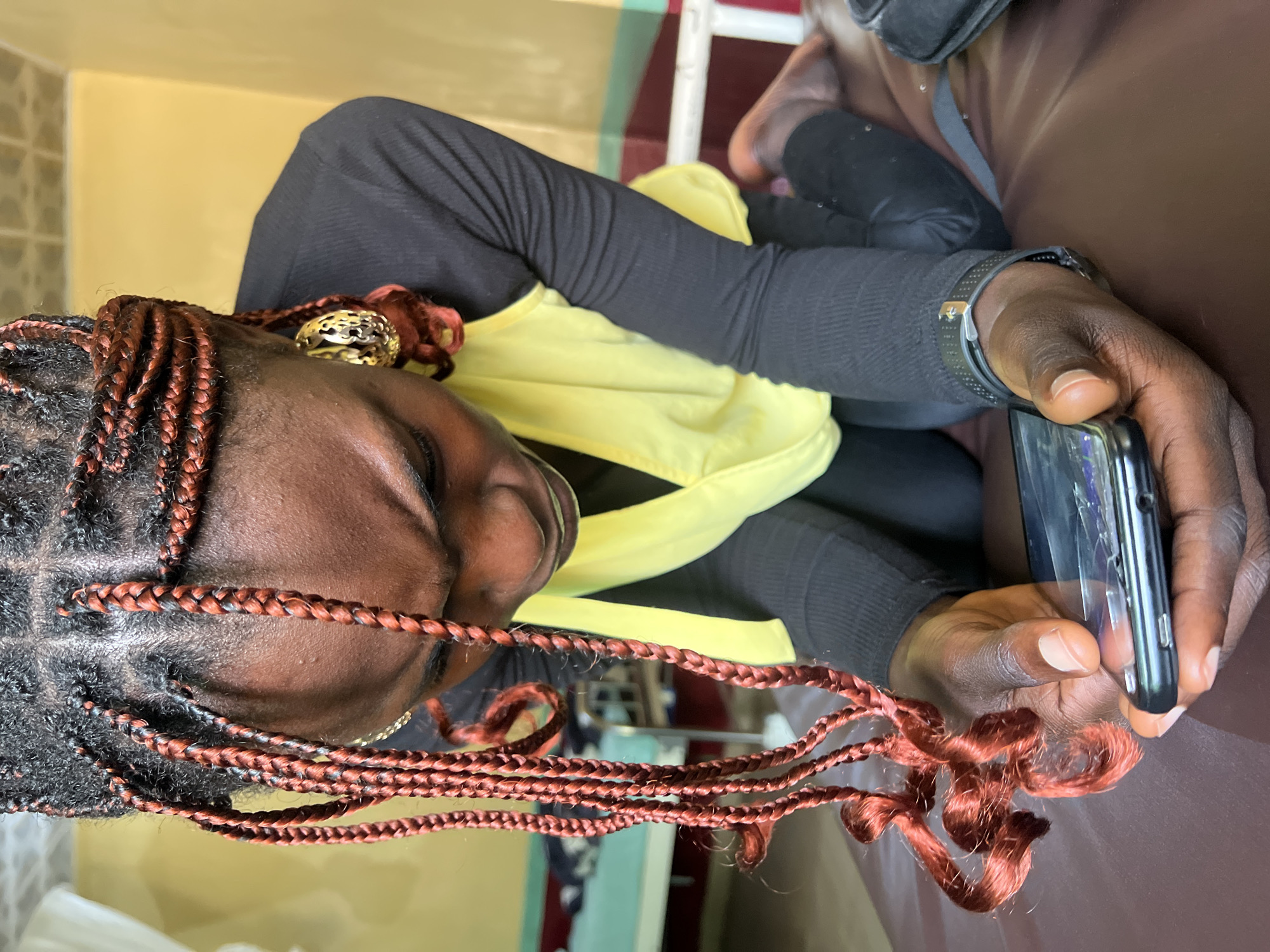 In Jos, Nigeria, a person with braided hair featuring red highlights kneels on a dark brown surface, intently looking at a smartphone held in their hands. The person is wearing a black long-sleeved shirt with a yellow sleeveless top over it. The background shows a room with walls painted in yellow and green stripes, and a white bed frame with a green mattress is visible. The scene appears to be indoors, possibly in a residential or medical setting.