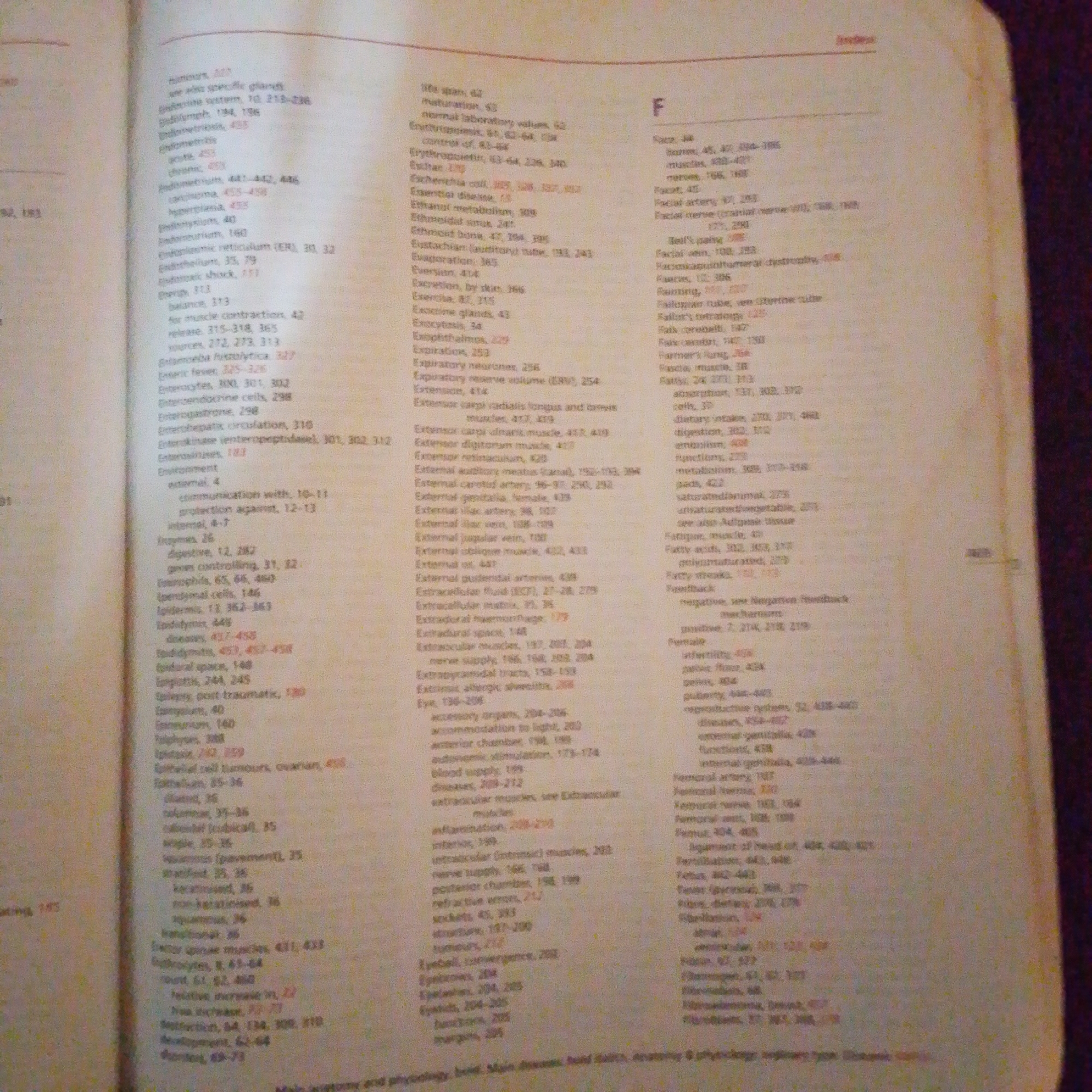 The image is a close-up, eye-level, slightly angled shot of an open book, specifically an index or glossary section, with text printed in columns on both pages. The lighting is uneven, with the left page being better illuminated than the right page. The page content is medical or biological terminology with corresponding page numbers. The book appears to be worn, with a slight yellowing of the pages, suggesting it's an older publication. The background is out of focus and shows a textured surface, likely a desk or table. There are no people, city of Jalingo, Nigeria, or any other external elements depicted in this image.