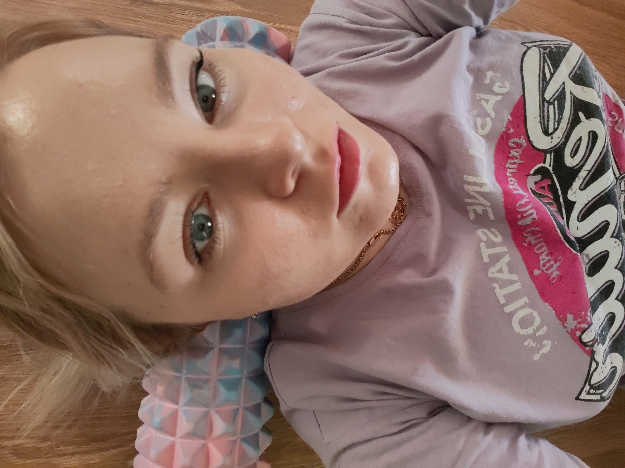 A woman is lying on her back on a wooden floor. She has light skin, blue eyes, and light blonde hair. She is wearing a lavender t-shirt with a graphic design. A textured blue and pink foam roller is positioned under her neck. A thin gold chain is visible around her neck. The location is unknown, and there is no indication that the image was taken in Izhevsk, Russia.