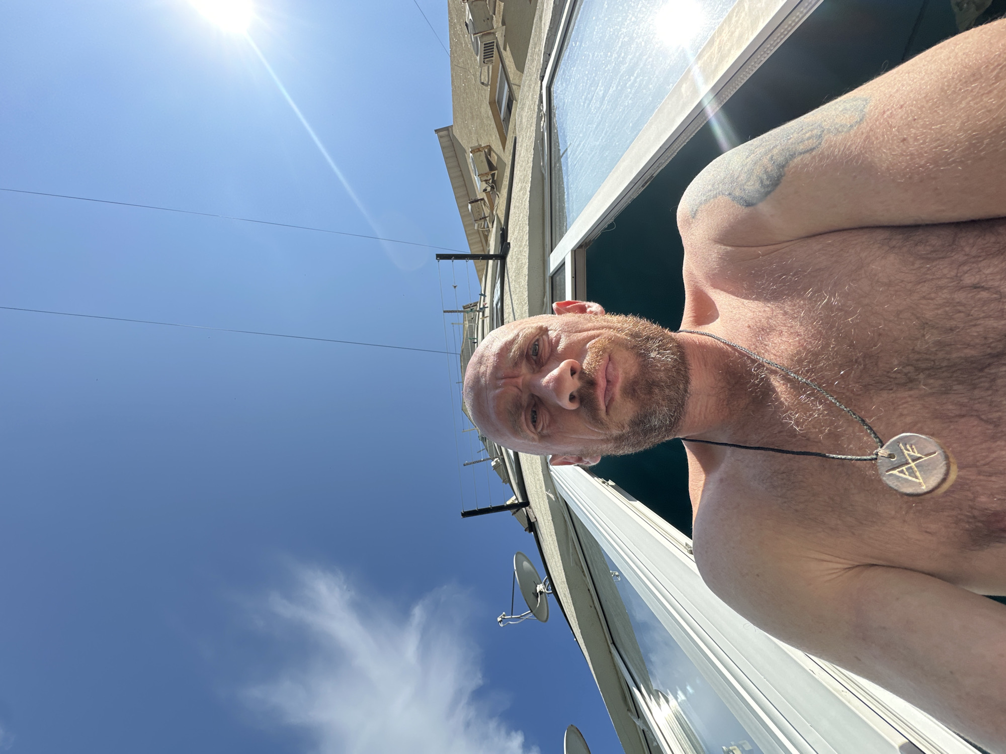 A low-angle, extreme wide shot captures a shirtless man with a bald head and a beard against a bright blue sky. He is positioned centrally in the frame, looking directly at the camera with a neutral expression. A circular pendant with an emblem hangs from a black cord around his neck. His left arm, visible to the right, has a tattoo. The man appears to be on a balcony or in an open window, with a white window frame running across the lower part of the image. In the background, a large, curved building rises, with visible windows, air conditioning units, and satellite dishes on the roof. Power lines are strung across the sky, and the sun shines brightly in the upper right corner, creating a lens flare. The scene is likely outdoors, during the daytime. The architectural style of the building, particularly the curved facade and the presence of multiple satellite dishes, could suggest a location in Sochi, Russia, given the region's common architectural trends and infrastructure.