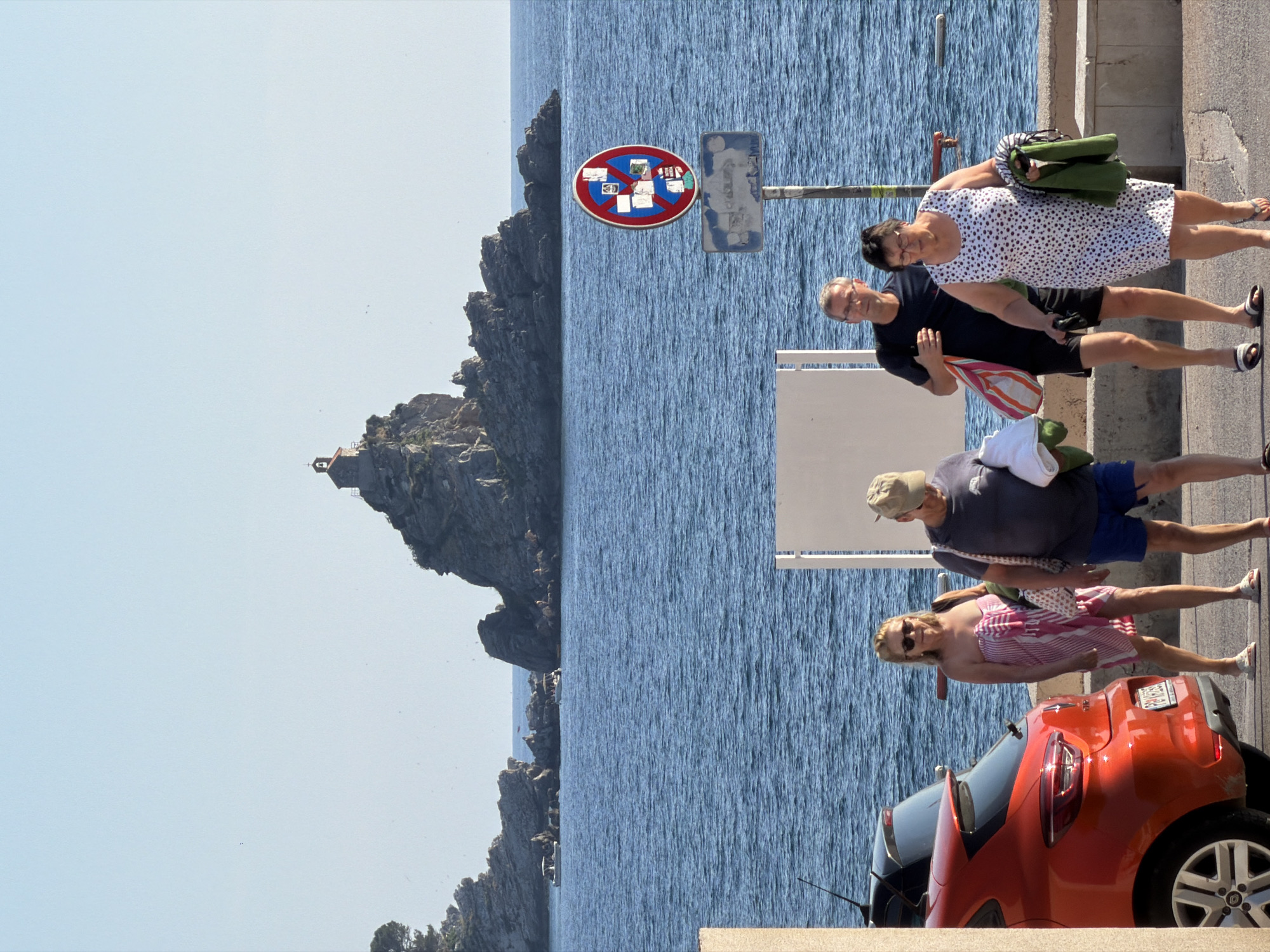 The image depicts a sunny day in Petrovac na Moru, Montenegro, with the iconic Church of the Holy Salvation perched atop a rocky islet in the background. In the foreground, a group of people are walking along a paved path by the sea. On the left, a bright orange car is parked. A woman in a pink and white striped towel is walking alongside a man wearing a baseball cap and shorts. Further to the right, a man in glasses and black shorts is walking, followed by a woman in a white polka-dot dress. The sea is a deep blue, with a few boats visible in the distance. A red and blue traffic sign with stickers on it is also visible on the right side of the image. The overall scene suggests a relaxed atmosphere on a summer day.