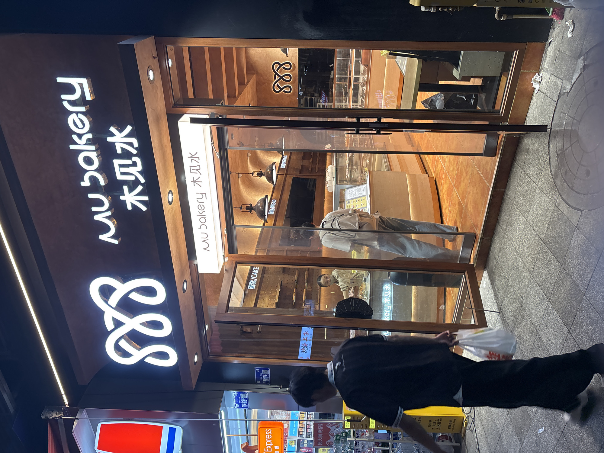 The image shows the storefront of "Mu Bakery" located in the Nan'an District, Chongqing, China. The bakery's name is written in both English and Chinese. The facade features a neon sign with the bakery's logo and name. The storefront has large glass windows and a partially open door, revealing the interior, including display cases and customers. A person in a black outfit is walking away from the bakery, carrying a white plastic bag. Along the street, there's a small retail area with signs for "Express" and other businesses.