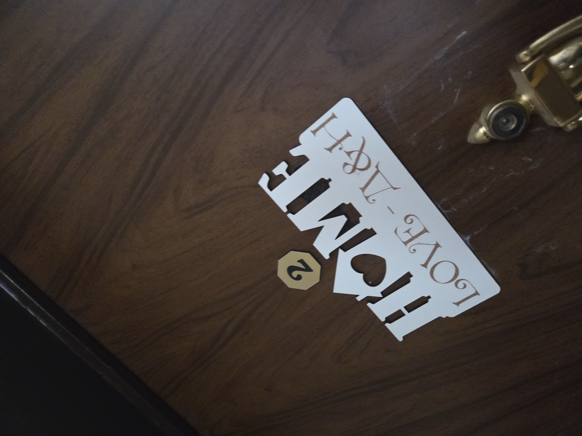 A close-up, slightly angled shot captures a dark brown wooden door or surface, exhibiting visible grain. In the center, a white decorative sign is affixed to the door. The sign features cut-out letters forming the word "HOME," where the 'O' is replaced by a heart shape. Above "HOME," the sign reads "LOVE - H&A" in a serif font. A small, golden, octagonal plaque with the black number '2' is placed below the "HO" of "HOME". To the upper right of the sign, part of a golden door knocker or peephole assembly is visible. The door's surface shows some faint white marks, possibly dust or scuffs. The image appears to be a detail of a residential or hotel room door, possibly indicating a room number and a personalized welcome. This scene is located in Pleven, Bulgaria.