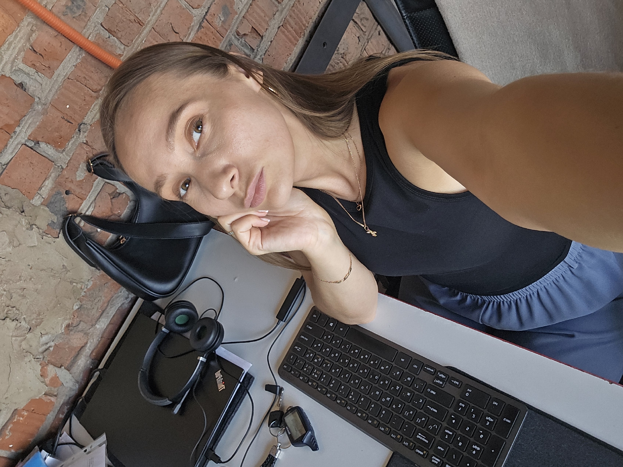The image shows a woman taking a selfie while sitting at a desk outdoors. She is resting her head on her hand, with a thoughtful or bored expression. She is wearing a black tank top and blue pants. On the desk are a keyboard, headphones, a laptop, keys with a remote, and a black handbag. The background shows a brick wall, suggesting an outdoor or industrial setting. The lighting suggests it is daytime. Visible text includes "bebinint" on the laptop and some Cyrillic characters on the keyboard. The overall impression is of someone working or studying in an unconventional environment. The location context provided is Chaikovsky, Russia.