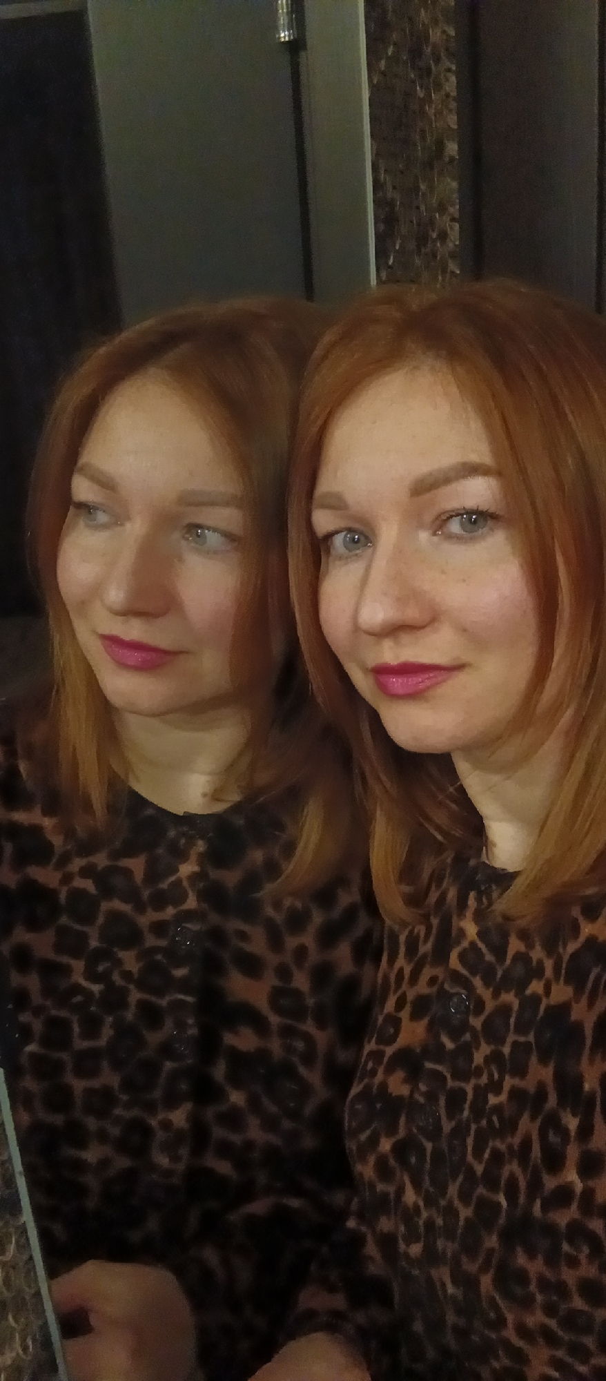 The image shows a woman with reddish-blonde hair and blue eyes, wearing a leopard-print top. She is wearing bright pink lipstick. The image seems to be a reflection in a mirror, as there is a second image of the same woman to the left, slightly out of focus and looking to the left. The background behind the woman consists of a dark gray wall to the left and a textured wall to the right. The location is Unknown, Unknown.