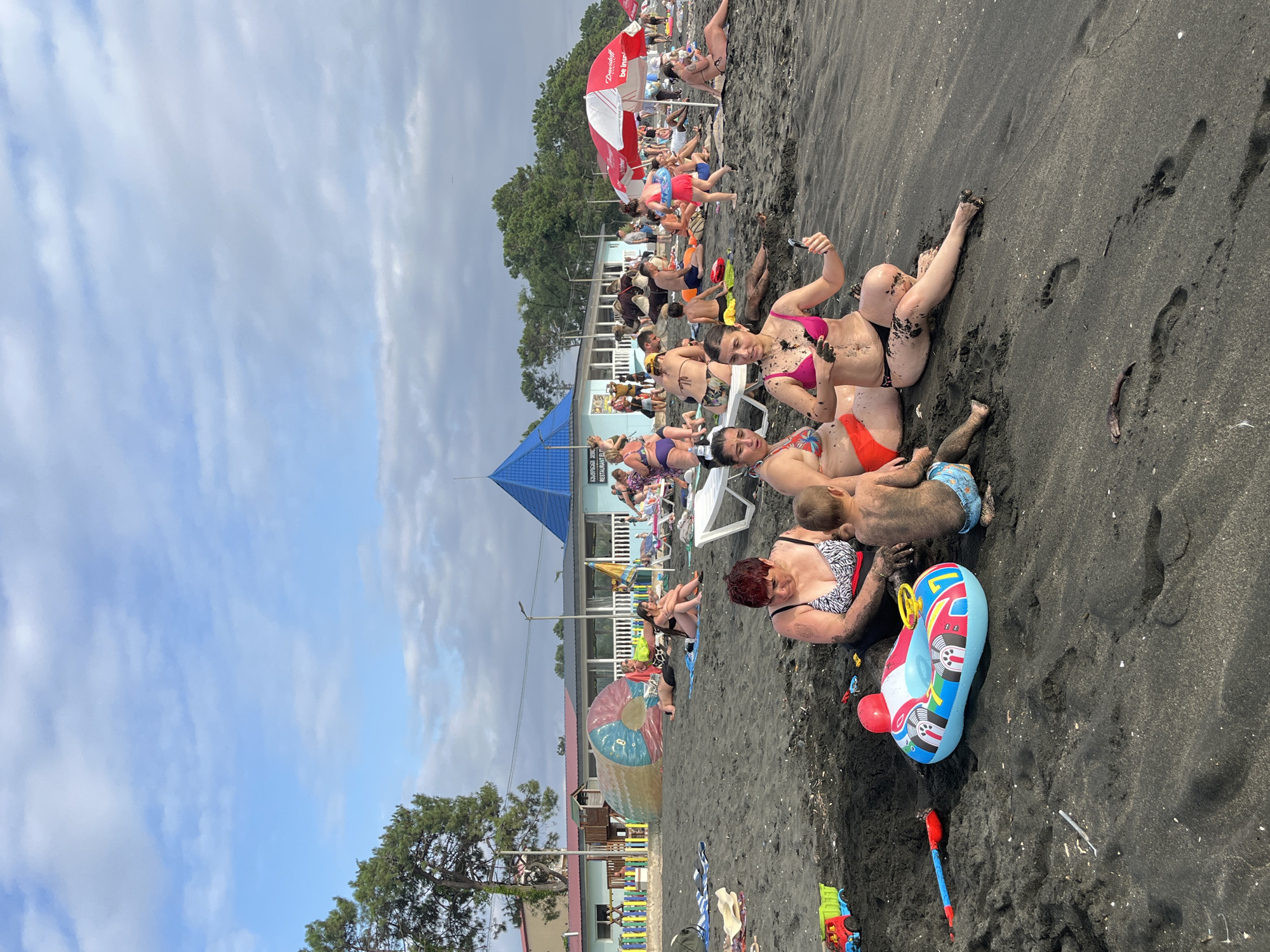 This image depicts a crowded black sand beach in Ureki, Georgia. The scene is filled with people of various ages, many of whom are wearing swimwear and relaxing on the sand or in beach chairs. Some individuals are tanning, while others appear to be sunbathing. A group of people, including women and children, are gathered closer to the camera, some covered in sand. One woman in a pink bikini is taking a selfie. A child is playing in the sand with a woman. There are several beach umbrellas providing shade, and a blue, pyramid-shaped structure stands out in the background. Behind the beachgoers, there are buildings that appear to be cafes or restaurants, with signs visible. A large inflatable toy, possibly a swimming ring, is on the sand near the foreground. Children's toys, including a red shovel and a toy truck, are also scattered on the sand. The sky is cloudy, suggesting it might be an overcast day, but the bright lighting implies daylight. The presence of trees in the background and the dark, fine sand are characteristic of the Ureki region. The overall atmosphere seems relaxed and leisurely, with people enjoying a day at the beach. One visible sign appears to say "Georgian Restaurant".