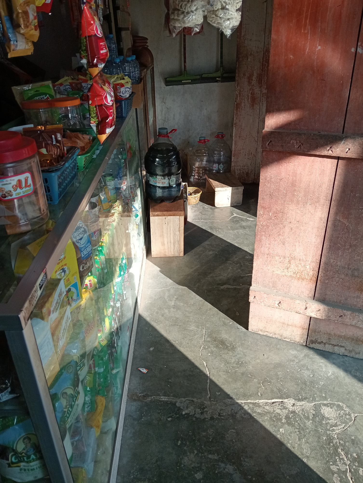 The image shows the interior of a small shop, likely a warung, in Grabag, Indonesia. The scene is illuminated by sunlight streaming in from an unseen window, casting strong shadows across the concrete floor and the various items displayed. To the left, a glass display case is filled with packaged snacks and drinks. Some of these packages are brightly colored, featuring text in what appears to be Indonesian. A large, clear plastic jar labeled "AL" with "RP 1000" on it is prominent in the foreground on the left. Behind this, a green plastic basket holds more packaged goods.
In the center of the image, on the floor, are several large plastic water bottles. One large dark blue bottle with a label that reads "Le Minerale" stands on a small wooden crate. Next to it, on a lighter colored wooden box, are two clear plastic water bottles, each with red caps. Another wooden crate is visible to the left of these water bottles.
To the right of the water bottles, a large, weathered, reddish-brown wooden door takes up a significant portion of the frame. The door has visible vertical planks and is fastened with simple metal hinges and what appear to be nails or screws. The wall behind the display case and the bottles is made of a light-colored, textured material, possibly concrete or plaster.
Hanging from the ceiling in the background are bags of white, puffy snacks, likely crackers or chips, in clear plastic packaging. Various shelves and racks holding more products are visible behind the display case. A green broom is leaning against the wall to the right of the hanging snacks. The overall impression is that of a modest, local convenience store stocked with common household goods and food items.