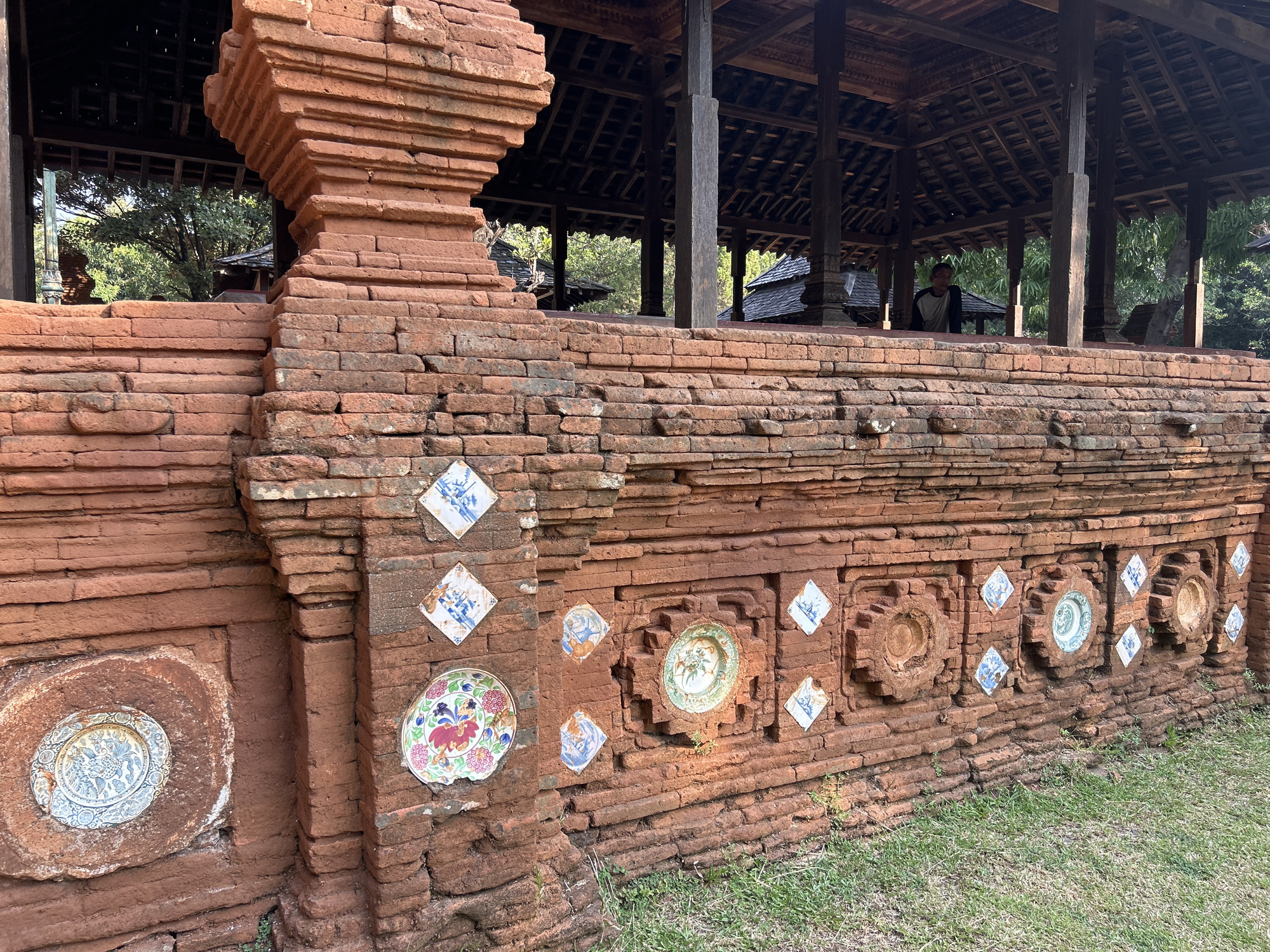 The image displays the exterior of a historical brick structure, likely part of a building or monument, in Cirebon, Indonesia. The structure is made of reddish-brown bricks and is adorned with various ceramic plates and tiles embedded within its walls. These decorations include patterned blue and white diamond-shaped tiles and larger, circular ceramic plates featuring floral and avian motifs. The architecture suggests a traditional style, with a covered wooden structure visible in the background, supported by wooden pillars. A person, appearing to be a male, is seated within this covered area, observing the surroundings. The ground in front of the brick structure is covered with green grass, indicating an outdoor setting. The overall scene conveys a sense of historical and cultural significance, typical of heritage sites.