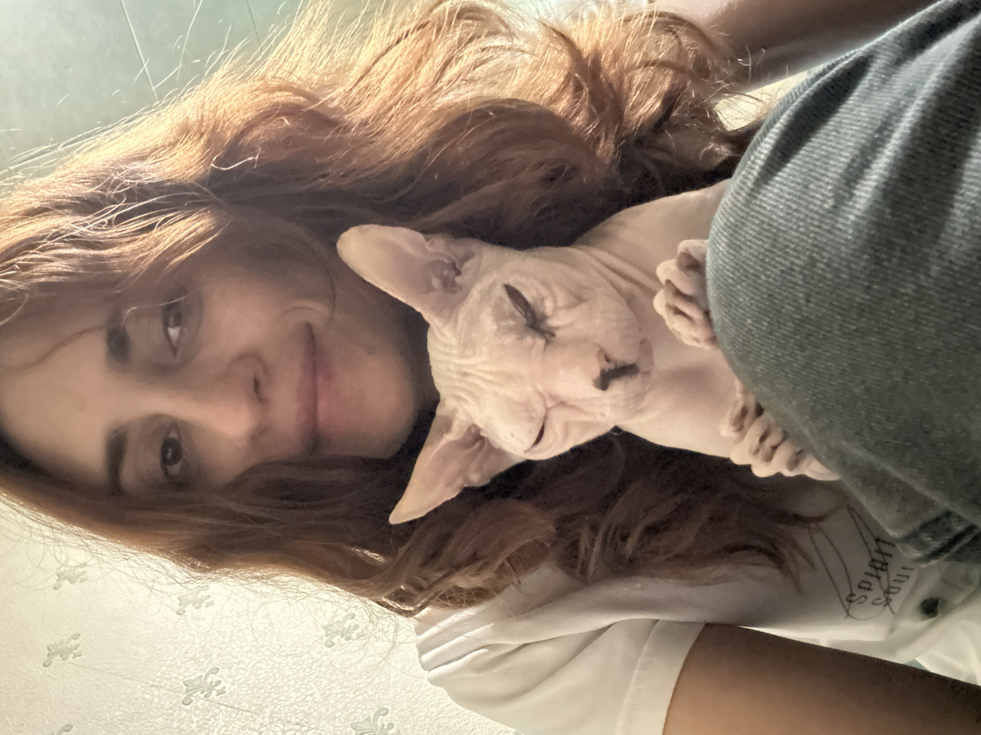 The image is a close-up selfie of a woman with reddish-brown, wavy hair and a Sphynx cat. The woman is wearing a white t-shirt and blue jeans, and is holding the cat in her lap. The cat, with its characteristic hairless skin and large ears, is looking down with its eyes closed. The background appears to be an indoor setting with wallpaper. The lighting suggests it may be daytime, possibly near a window. There are no recognizable landmarks or specific details that would indicate the location is Togliatti, Russia.