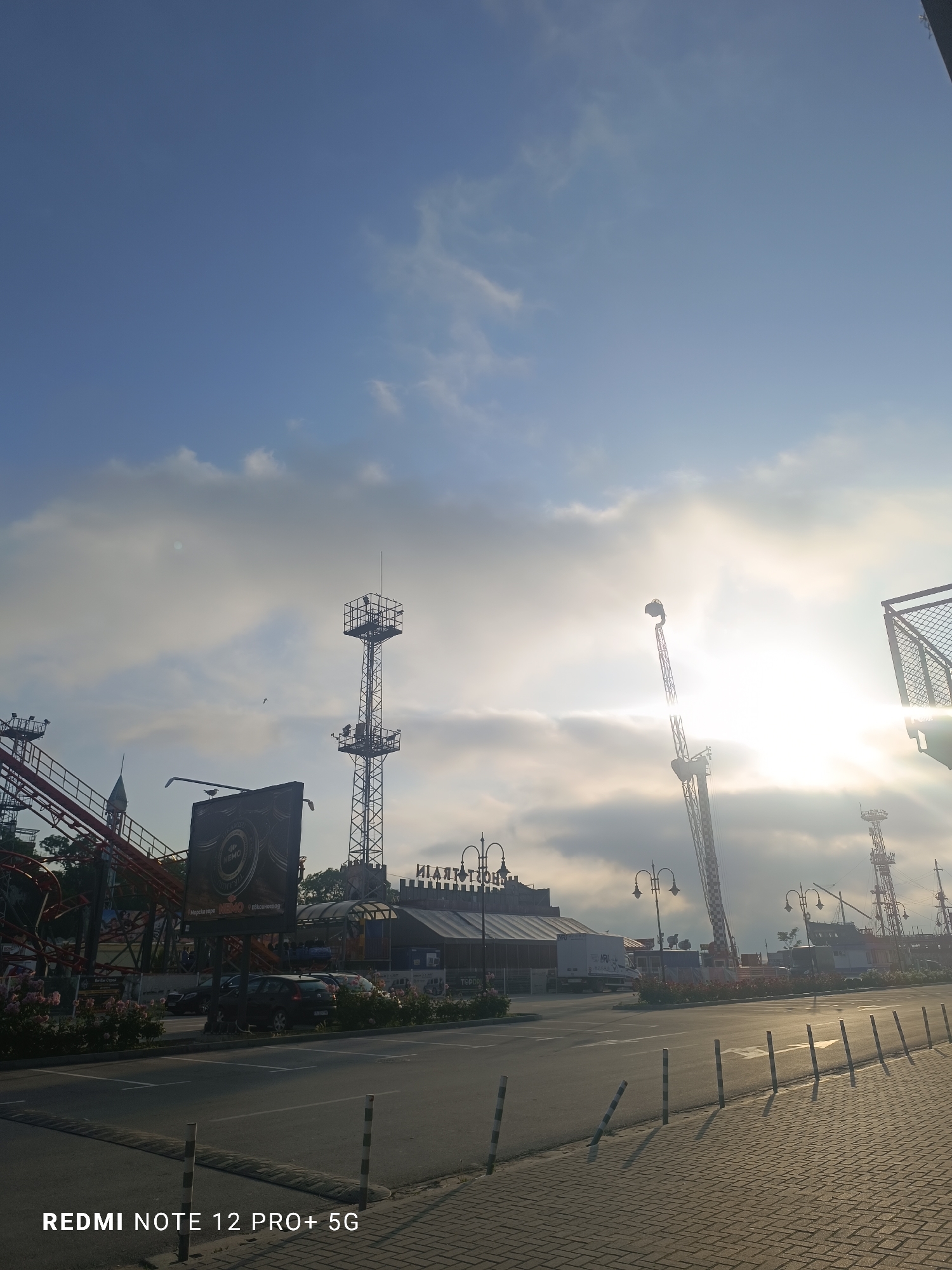 The image captures an outdoor scene at what appears to be an amusement park or entertainment complex in Varna, Bulgaria, during either sunrise or sunset, indicated by the low sun casting long shadows and creating lens flare on the right. The sky is partly cloudy with patches of blue and hazy light filtering through the clouds.
In the foreground, a paved road or parking area is visible, bordered by a brick sidewalk and a line of striped bollards. Bushes with pink roses line the road. In the midground, red roller coaster tracks are prominent on the left. Several tall metal structures, resembling parts of thrill rides like drop towers, are scattered across the frame, silhouetted against the bright sky.
A large billboard stands in the midground, displaying the word "NEMO" and the Cyrillic text "Морско око" (Sea Eye). A bird is perched on top of this billboard. Behind it, a building features the reversed Cyrillic text "ЦИРК МОРСКО ОКО" (CIRCUS SEA EYE), suggesting it might be a circus or an attraction with a marine theme. Another building with a castle-like facade is also discernible. A black car is parked near the buildings.
The scene is devoid of people, creating a quiet and still atmosphere. The camera watermark "REDMI NOTE 12 PRO+ 5G" is visible in the bottom left corner.