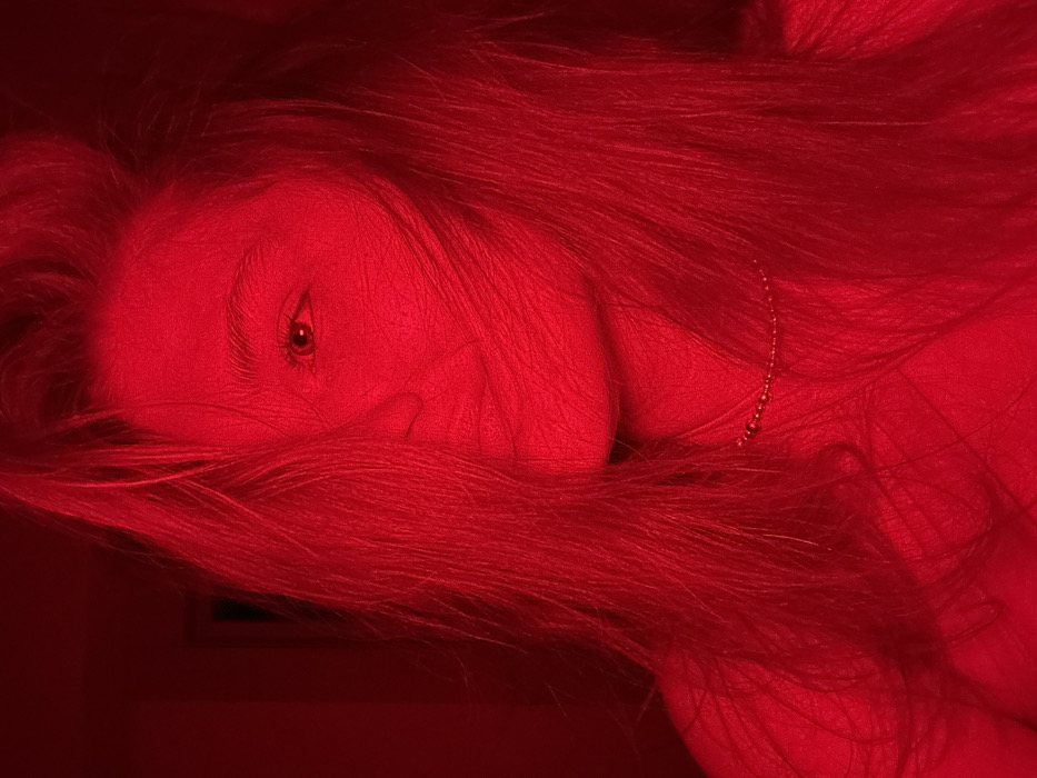 An image, captured in the city of Noviy, Russia, displays a close-up of an individual, likely female, illuminated predominantly by red light. The subject's face is partially obscured by long, light-colored hair, which covers the left side of her face. Her right eye is visible and directed towards the camera. A beaded necklace is worn around her neck. The background is indistinct and dark, indicating an indoor setting under low light conditions where a strong red light source is dominant. No specific actions or interactions beyond posing are discernible.