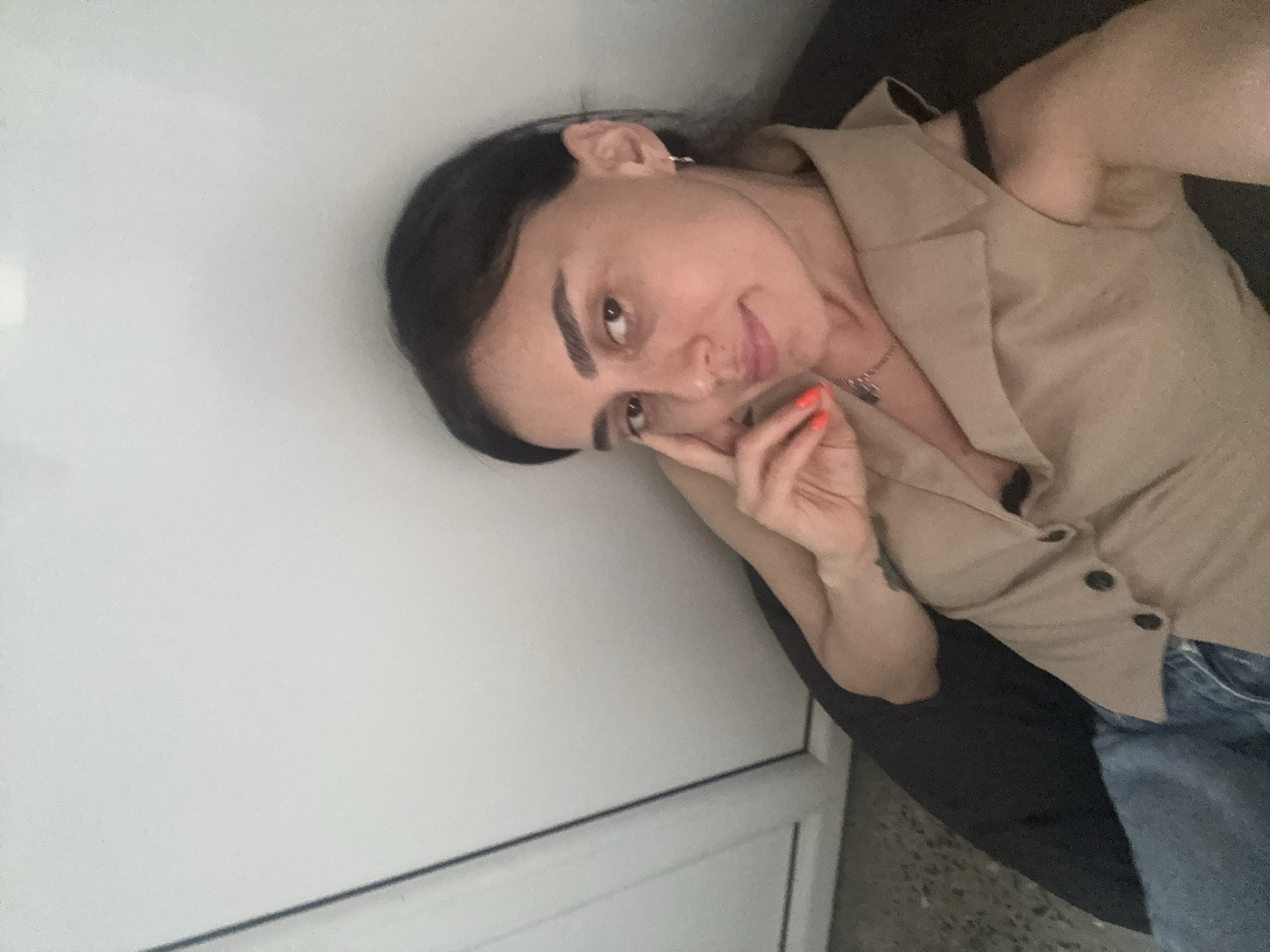 In this selfie taken indoors, a woman with dark hair is partially visible, leaning against a dark surface. She is wearing a light brown sleeveless vest over a dark top and blue jeans. Her right hand is raised to her face, with bright orange nail polish visible on her fingertips. She is wearing a small silver hoop earring in her right ear. The background consists of a plain, light-colored wall and a portion of a white door or partition. There is no indication of the location being specifically in Tbilisi, Georgia.