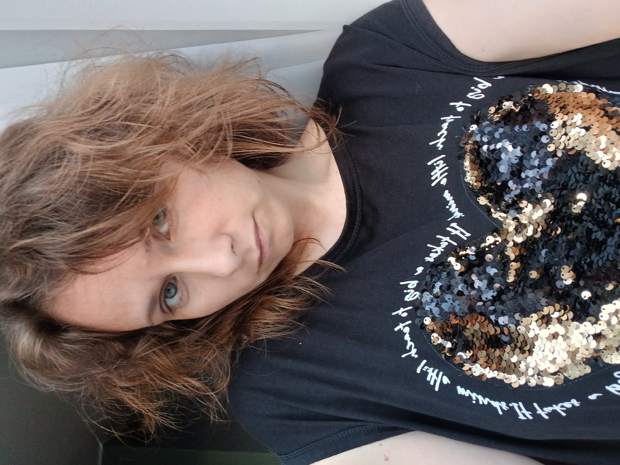 A medium shot, taken from a slightly low angle, shows a woman with light brown, messy hair. She is wearing a black t-shirt with a large heart design made of black and gold sequins. White cursive text encircles the heart. The woman is looking directly at the camera with a neutral expression, her mouth slightly open. Her skin appears fair with some freckles. The background is blurred, with a dark area to the left and a light, sheer curtain to the right. There is no visible indication of the location being Yekaterinburg, Russia.