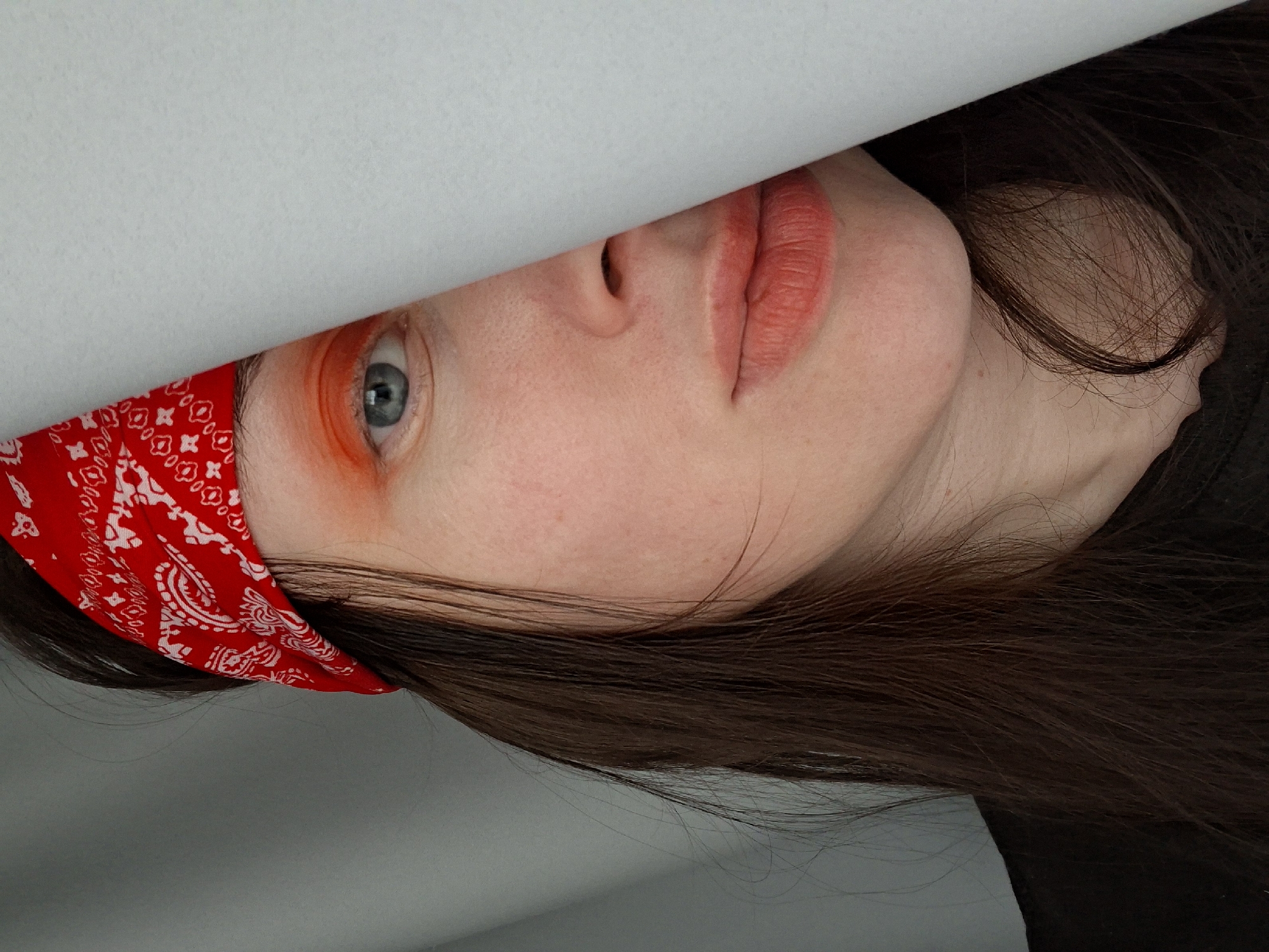 A portrait of a person with fair skin, blue eyes, and dark brown hair. The person is partially obscured by a sheet of white paper or fabric. The portion of the face visible includes the nose, lips, and part of the right eye. The right eye has orange makeup around it. The person is wearing a red bandana with a white pattern on their head. The environment is not specified.