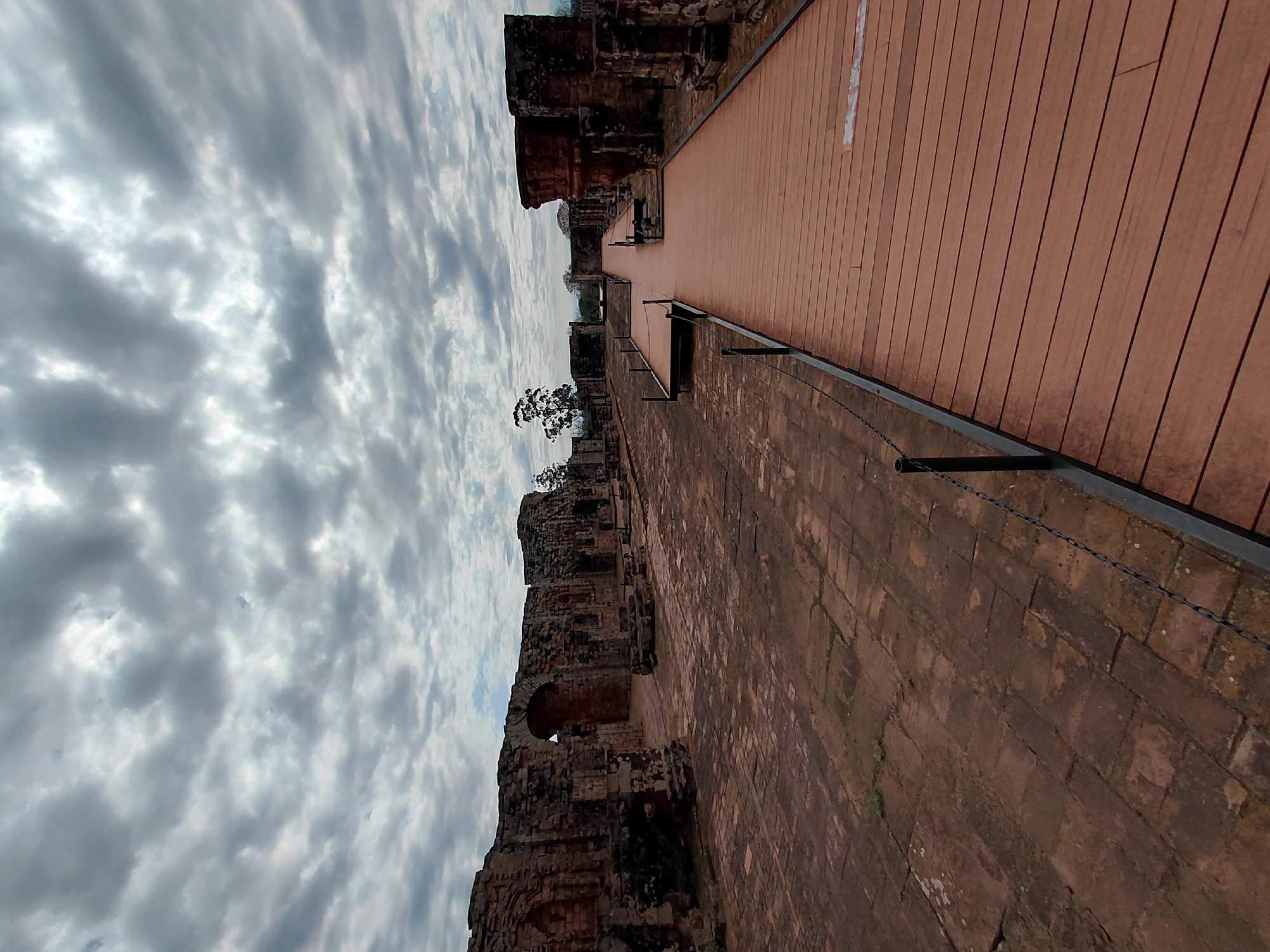 The image shows the ruins of the La Santísima Trinidad de Paraná mission, a UNESCO World Heritage site in Trinidad, Paraguay. The foreground features a raised wooden walkway with black metal railings. Beyond the walkway, ancient stone structures with arched doorways and weathered brickwork stand under a cloudy sky. The ground within the ruins is paved with reddish-brown bricks. The scene depicts a historical site, likely a former Jesuit mission, with preserved architectural remains. No people are visible in the image. The overall environment is one of historical significance and architectural preservation.