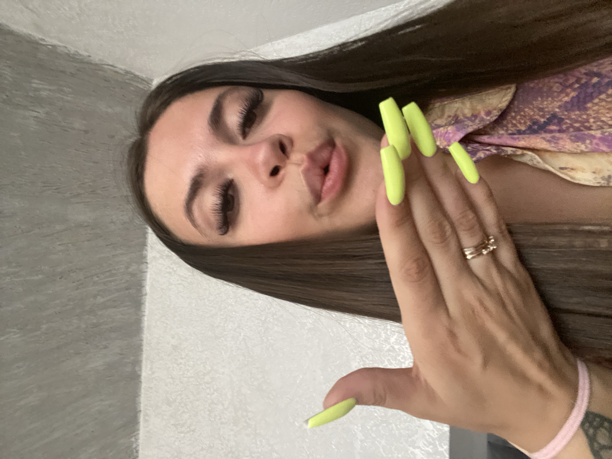 A woman with dark brown hair and long, bright green acrylic nails is looking at the camera with her lips pursed. She is wearing a light pink bracelet on her left wrist and a patterned shirt with shades of purple, gold, and black. A gold ring is visible on her left hand. The background consists of a textured white wall and a grey, textured ceiling. There is no indication of the location being Chalda, Russia.