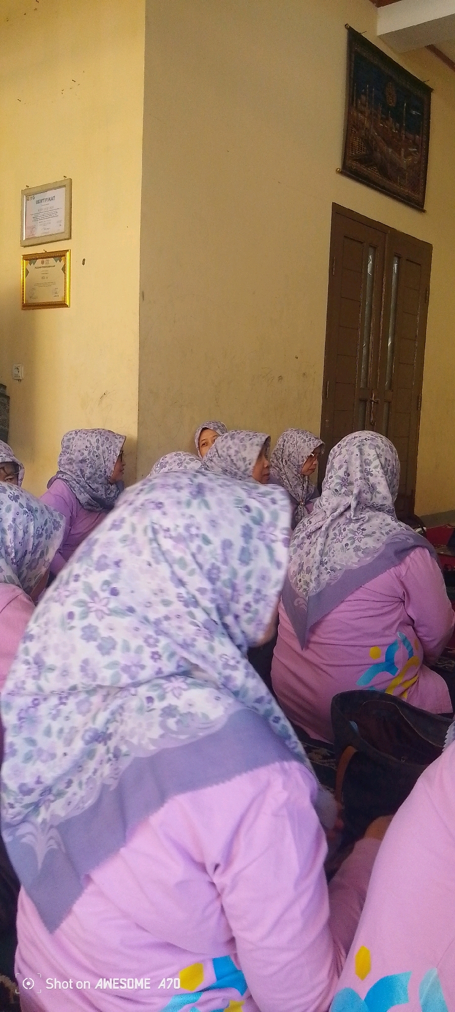 The image shows a group of women wearing headscarves and light pink outfits, possibly uniforms. They are seated on the floor, facing away from the camera or looking to the side, suggesting they are attending an event or gathering. Some of the women are looking at each other, indicating interaction within the group. The background features a light yellow wall with framed certificates hanging on it, and a dark wooden door to the right. A tapestry hangs on the wall above the door. The overall impression is that of a formal or semi-formal event taking place indoors, likely in an educational or community setting. There is no specific visual information that points to the location being the city of Bandung, Indonesia.