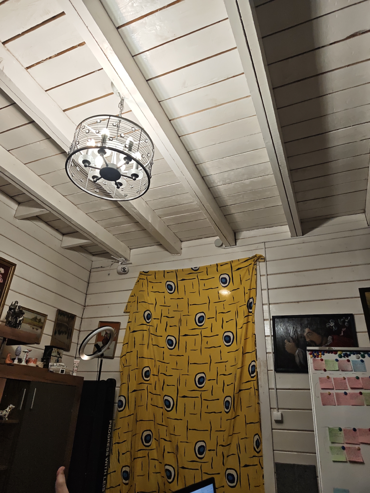 The image depicts an interior space with a focus on the ceiling and nearby furnishings. The ceiling is constructed of painted white planks with exposed beams. A modern, cage-style chandelier hangs from the ceiling. A yellow fabric with a repeated circular motif hangs against a wall of similar painted planks. To the left of the fabric, there is a display cabinet with various framed pictures and decorative objects. A ring light stands on a tripod near the cabinet. To the right of the yellow fabric, a whiteboard covered in colorful sticky notes is visible. The room has an unfinished aesthetic with exposed construction and a mix of contemporary and traditional decor elements. The location is Свистуха (кузяевский С/о), Russia.
