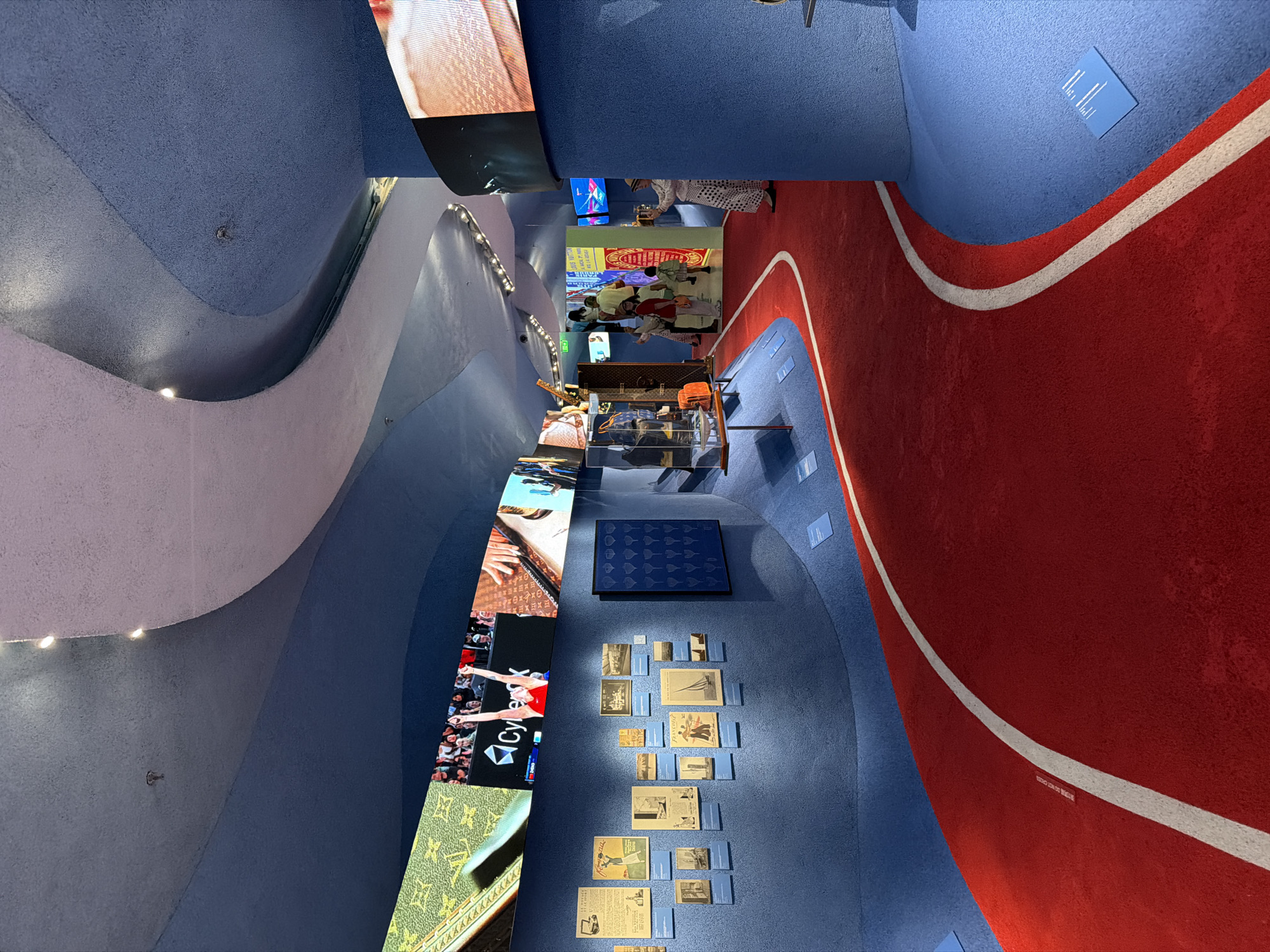 The image captures an interior view of a modern museum exhibit in the Jing'an District of China. The exhibition hall features a curved design with a blue textured ceiling and walls, bisected by a red carpeted pathway with a white stripe border.
On the left side of the frame, a series of large, vertical video screens display various images, including portraits, sports footage featuring an athlete with "Cyber" emblazoned on their uniform, and a pattern resembling Louis Vuitton's monogram.
Along the blue wall, there are a framed display of blue square tiles, photographs, and informational panels. People are seen in the background, interacting with the exhibits.
In the distance, individuals are visible in the exhibition space, looking at artifacts displayed in glass cases. A white-clad individual is seen on the right side.