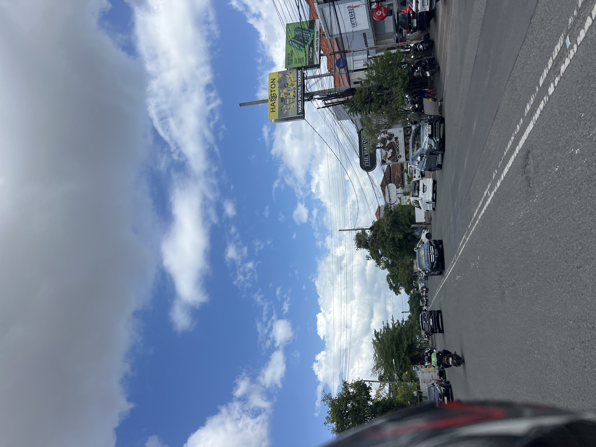 The image captures a street scene in Denpasar, Indonesia, under a partly cloudy sky. The perspective is from the viewpoint of someone on a motorcycle, looking forward along a paved road. Several vehicles, including cars, motorcycles, and a van, are present on the road, some of which appear to be moving. On the right side of the road, there are buildings and businesses, including a sign that reads "HASTON" with images of tools and "YANG PUTRA TEKNIK". Another sign on the right displays "INTEGRID". Trees line the sides of the road, and power lines are visible overhead. The environment appears to be an urban or suburban area with commercial activity.