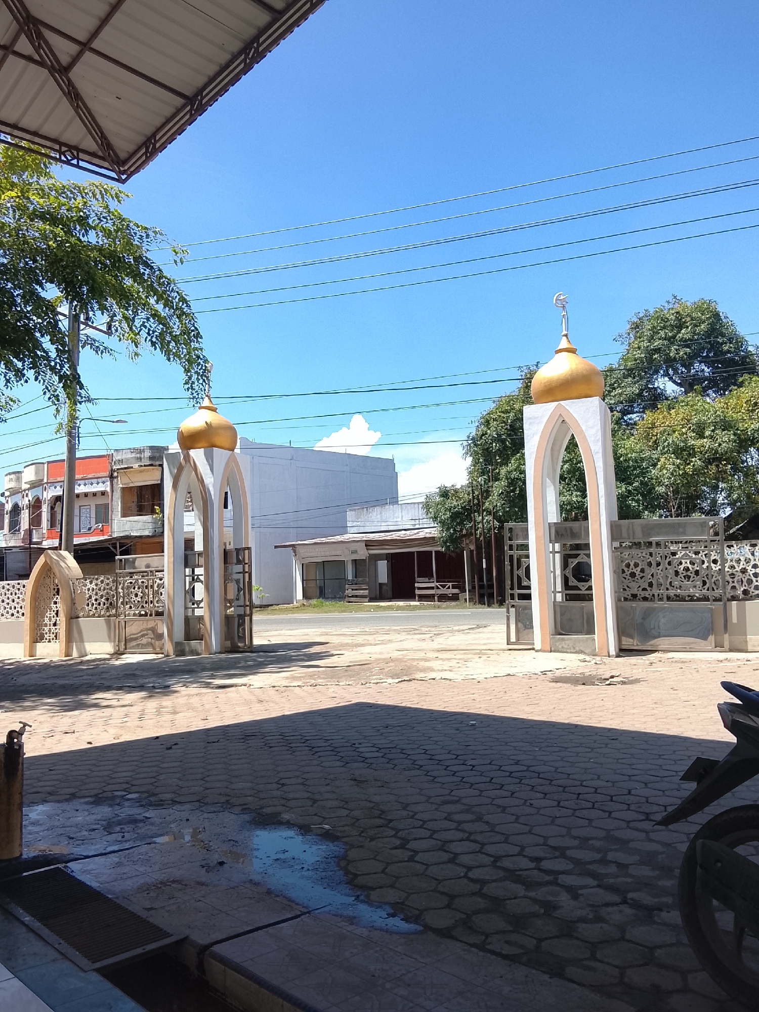 The image captures an outdoor scene, likely in the city of Meunasah Bie, Indonesia, under a clear blue sky. In the foreground, a paved area with a hexagonal pattern is partially wet, suggesting recent rain or cleaning. A metal drain grate is visible in the lower left. To the right, the rear section of a dark-colored motorcycle is visible.
Dominating the midground are two mosque gateways, characteristic of Islamic architecture. Both feature white archways with gold-colored domes topped with crescent moons. The gateway on the left is smaller and has an ornate, cut-out design. The gateway on the right is larger and more prominent, with a fence behind it featuring a geometric pattern. Beyond the gateways, a street is visible with some buildings lining it, including a white multi-story building and a smaller structure with a wooden roof and open sides. Trees with green foliage are scattered throughout the background. Overhead, a section of a metal roof structure extends from the upper left, and power lines stretch across the sky. The overall impression is a daytime scene in a built-up area with distinct Islamic architectural elements.