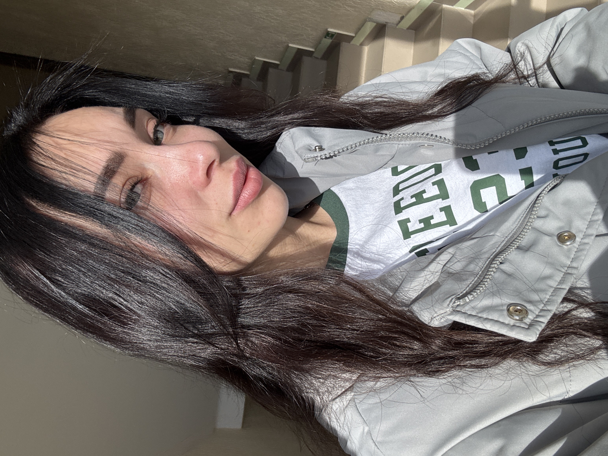 This portrait image, possibly a selfie, captures a young woman at an angle, rotated 90 degrees counter-clockwise. The scene is set in what appears to be an indoor or semi-sheltered outdoor location, possibly in Chaikovsky, Russia, with a light-colored staircase and railings visible in the background.
The primary subject is a woman with long, dark, wavy hair, fair skin, and light-colored eyes, possibly green or hazel. Her expression is neutral and relaxed. She is wearing a light gray or silver-colored quilted jacket with a visible zipper and snap buttons. Underneath, she has a white t-shirt featuring green text, which clearly reads "LEGENDS" and partially shows what could be "U" or "UNIVERSITY" after it.
The image is characterized by strong, direct sunlight, suggesting a clear, sunny day, likely mid-morning or afternoon. This harsh lighting creates distinct highlights on her hair and face, and shadows on her clothing and the background. Her dark hair is spread across her shoulders and the upper part of her jacket. The visible details include the texture of her hair, the quilted pattern and hardware (zipper, snap buttons) of her jacket, and the bold green lettering on her t-shirt. The overall mood conveyed is calm and sunlit.