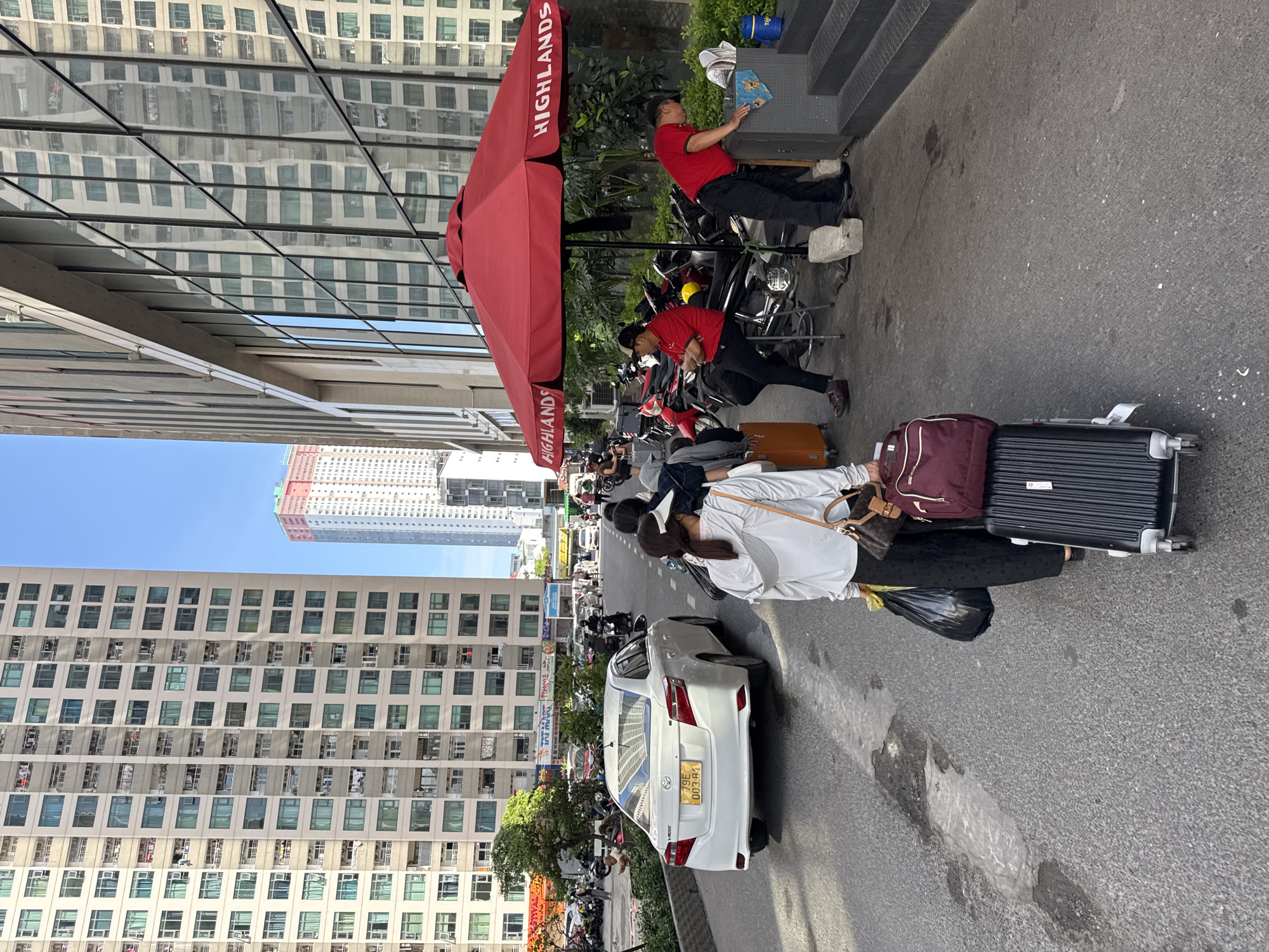 The image depicts a street scene in Bac Nha Trang Ward, Vietnam, likely in front of a hotel or large building. In the foreground, a woman with a white long-sleeved shirt and black pants is pulling a black rolling suitcase and carrying a maroon duffel bag on top of it. She also has a black trash bag in her left hand and a brown handbag slung over her right shoulder. Behind her, a white Toyota sedan with a yellow license plate is parked on the street. To the right, under a large red umbrella with "HIGHLANDS" printed on it, two men in red shirts are standing or sitting. One man is leaning over a grey cabinet, and the other is sitting on a stool. Numerous motorcycles are parked in the background. The buildings in the background are tall, multi-story structures, with one prominent building displaying reflections of other buildings in its glass facade. The environment appears to be an urban setting with a clear blue sky.