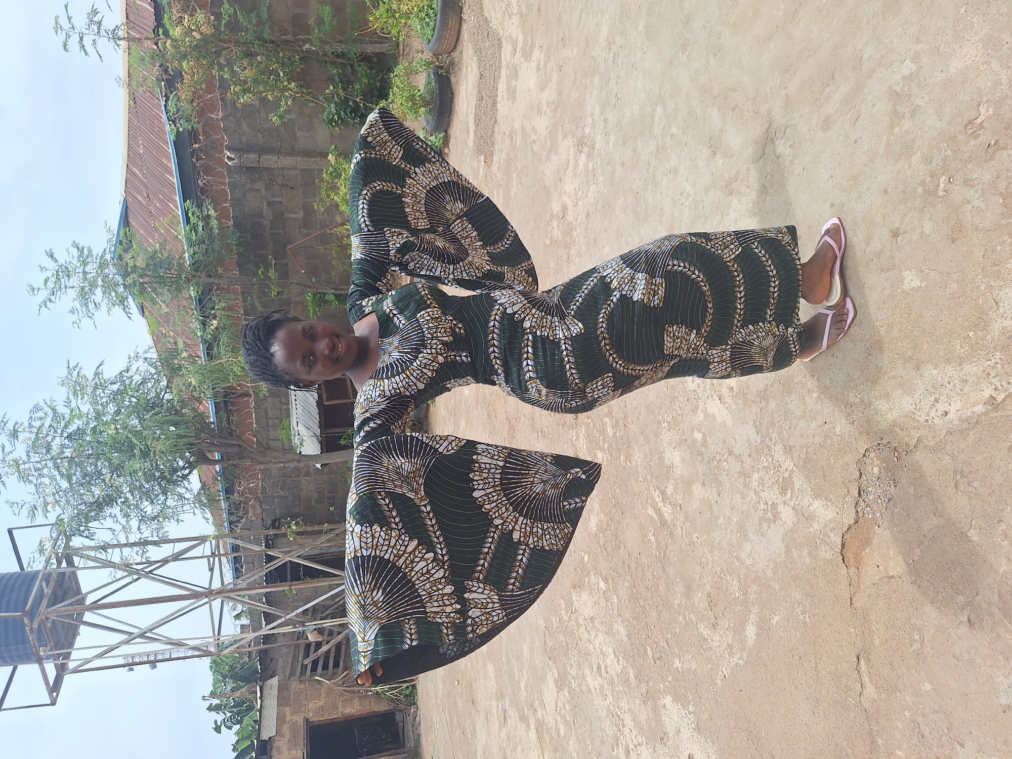 A woman stands on a concrete surface in Sabon Tasha, Nigeria. She is wearing a long, patterned dress with flared sleeves and pink flip-flops. Behind her, there is a water tower, trees, and a building with a corrugated metal roof.
