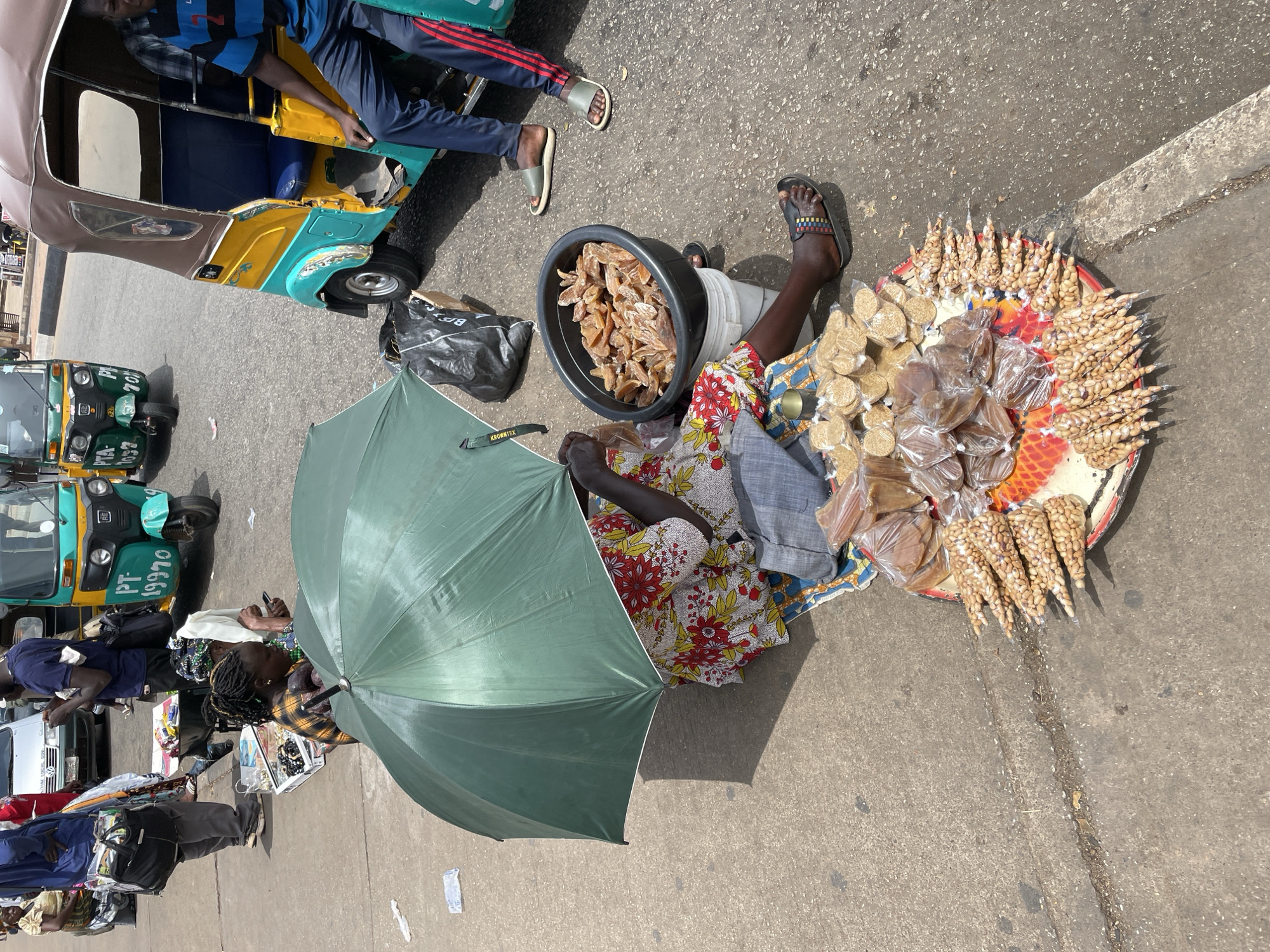 The scene is a street in Barakin Lerawi, Nigeria. A woman sits on the ground next to a curb, using a green umbrella for shade. She sells various foods, including dried fruit in a black bowl and packaged peanuts arranged on a circular tray. Beside her, two people sit in a three-wheeled vehicle. Several other three-wheeled vehicles are parked in the background. The area appears to be a busy street.